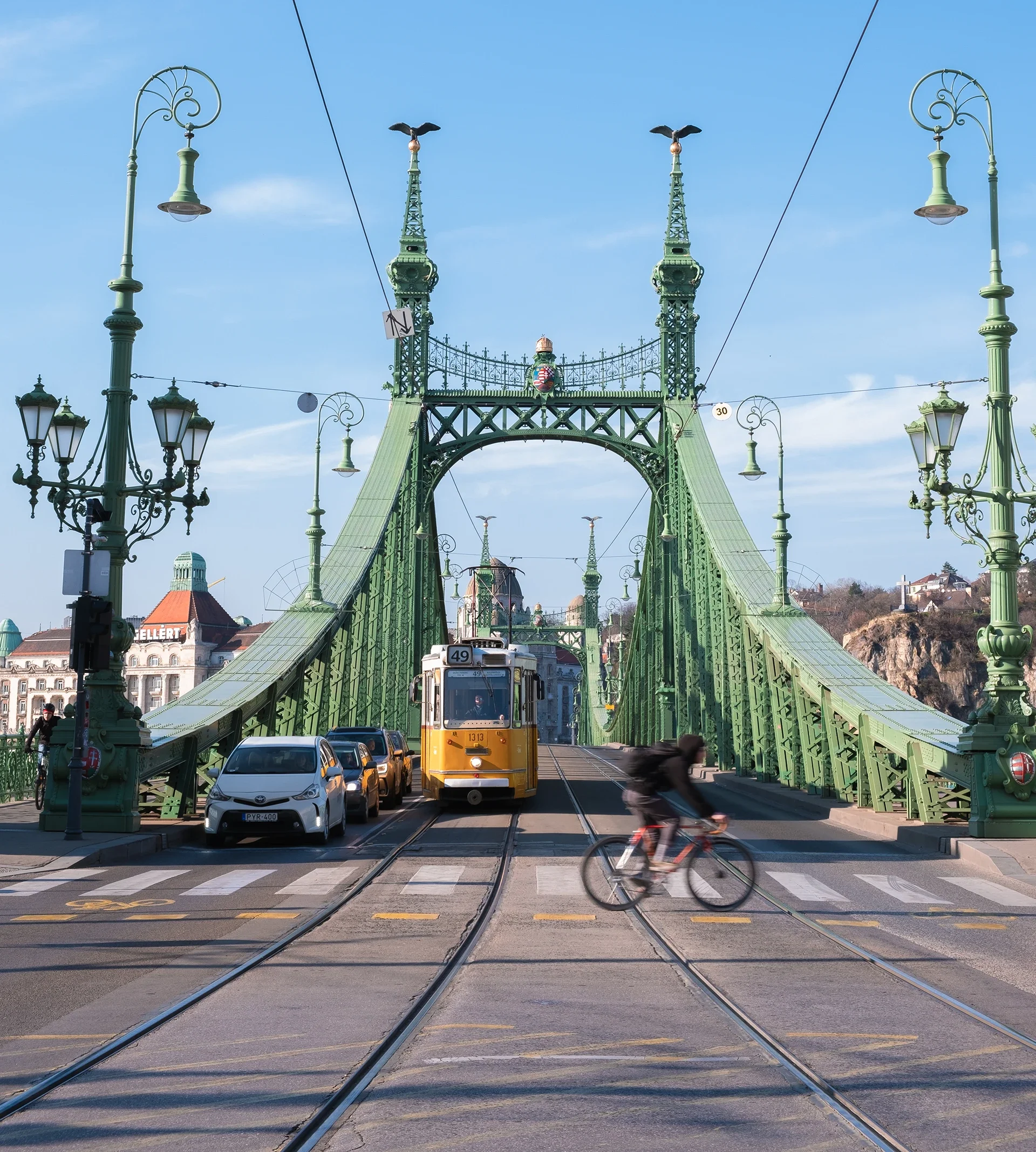 View of the Liberty Bridge in Budapest, Hungary, featuring green ornate iron architecture, a tram, cars, and a cyclist crossing over the bridge with a clear blue sky in the background.