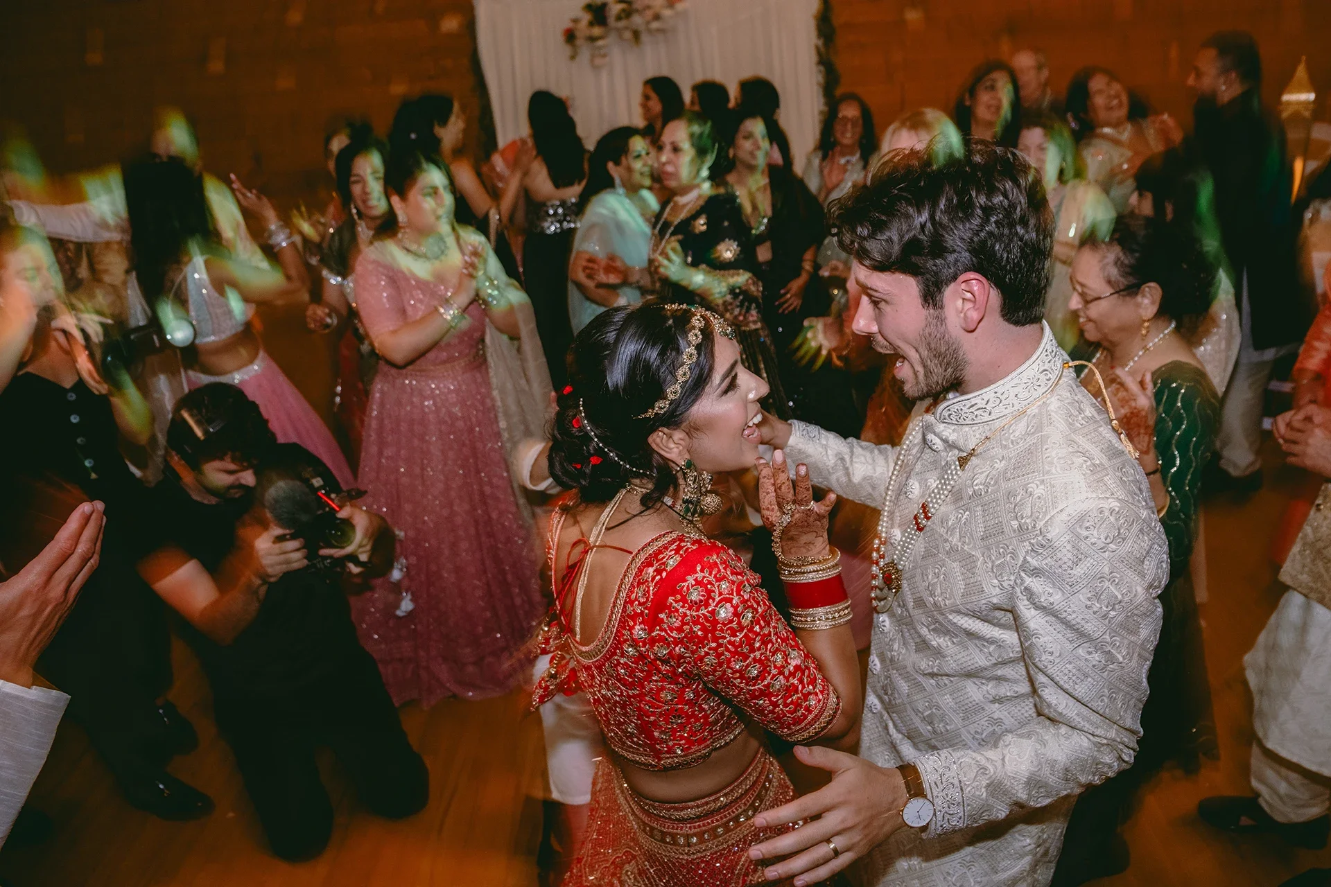 A couple dancing closely at a celebration, surrounded by well-dressed guests in colorful traditional attire, with a photographer capturing the moment.