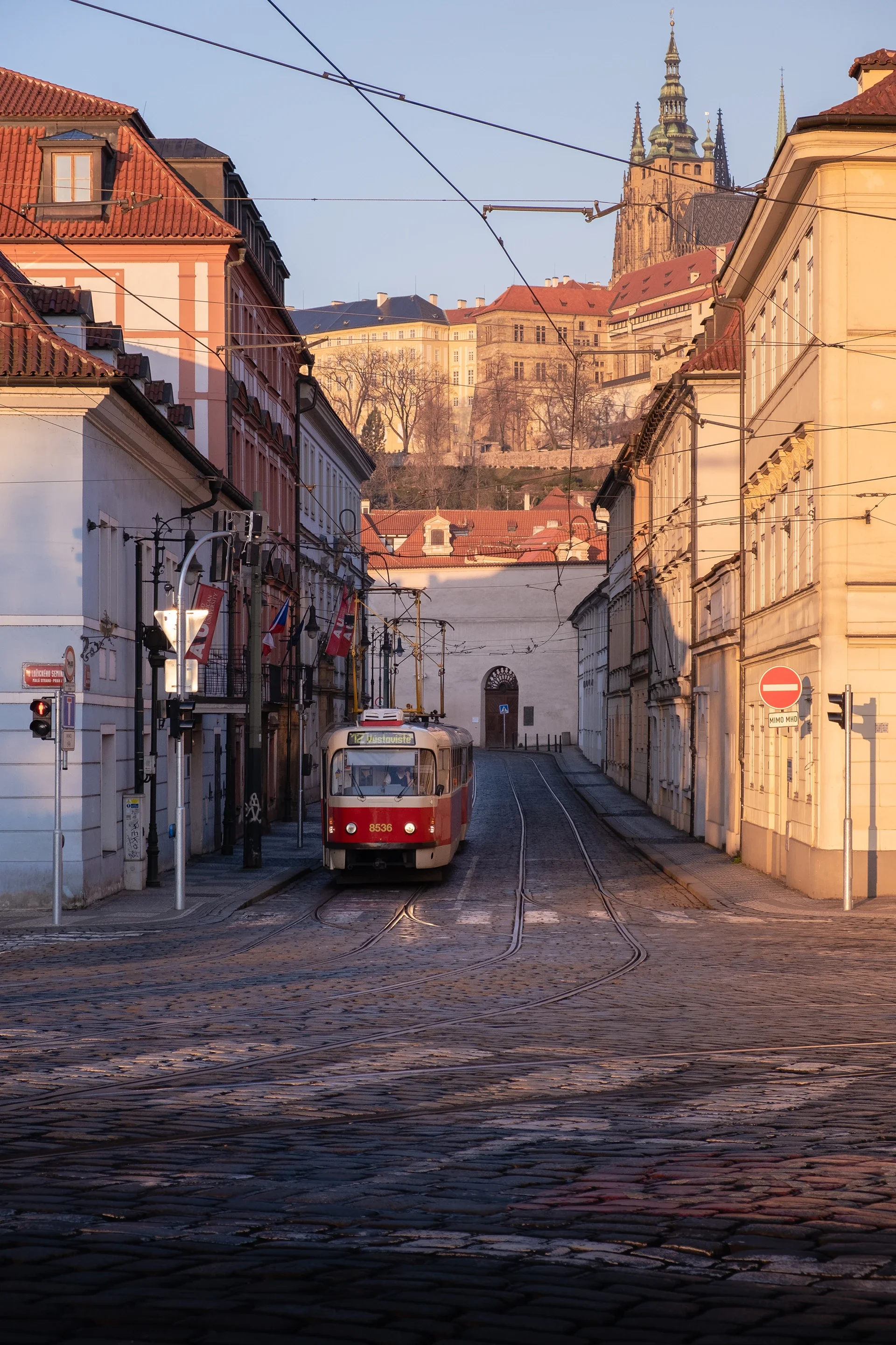 A tram traveling along cobblestone street, with buildings on either side and a castle on a hill in the background, during sunset.