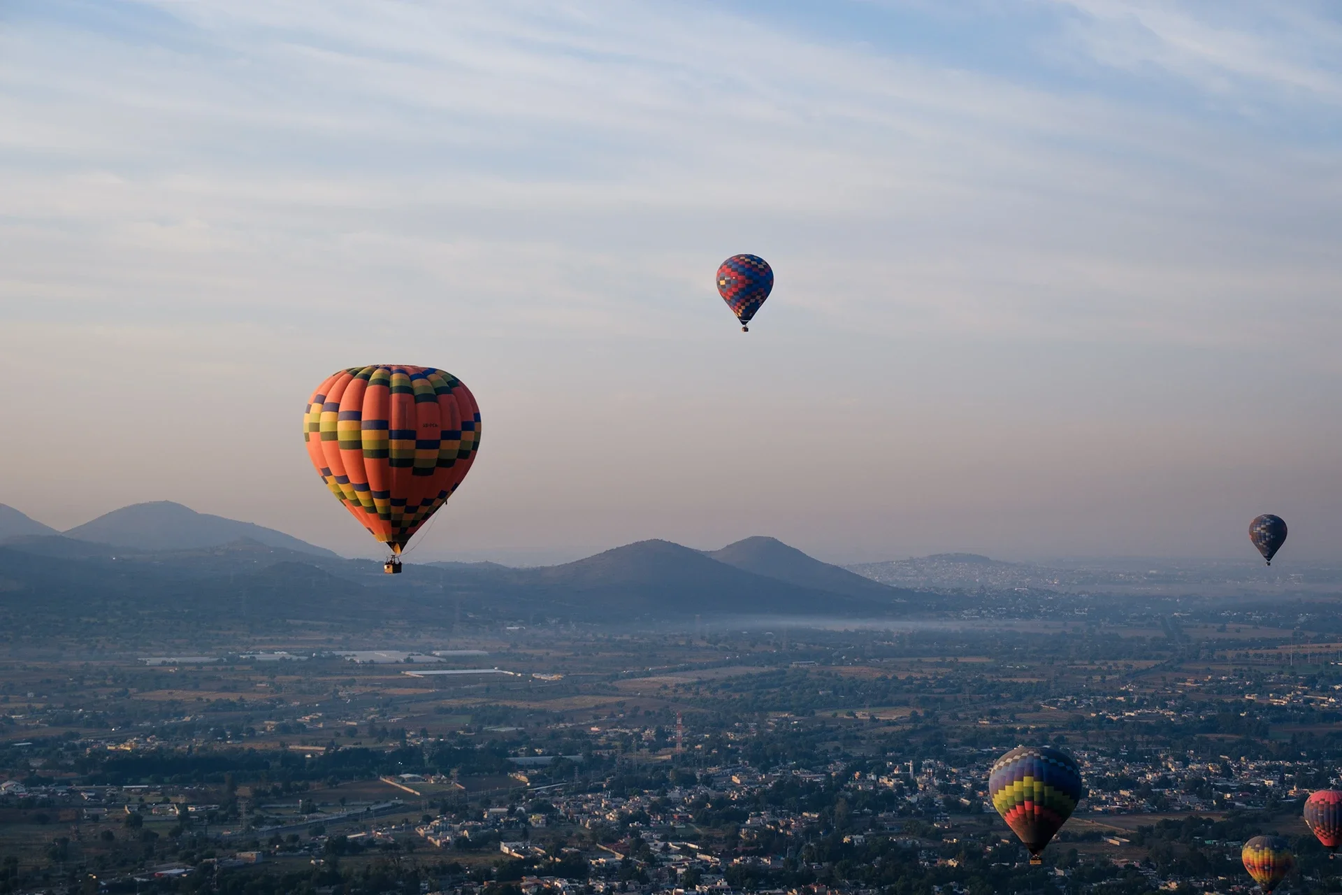 Several colorful hot air balloons floating over a city and mountainous landscape under a cloudy sky.