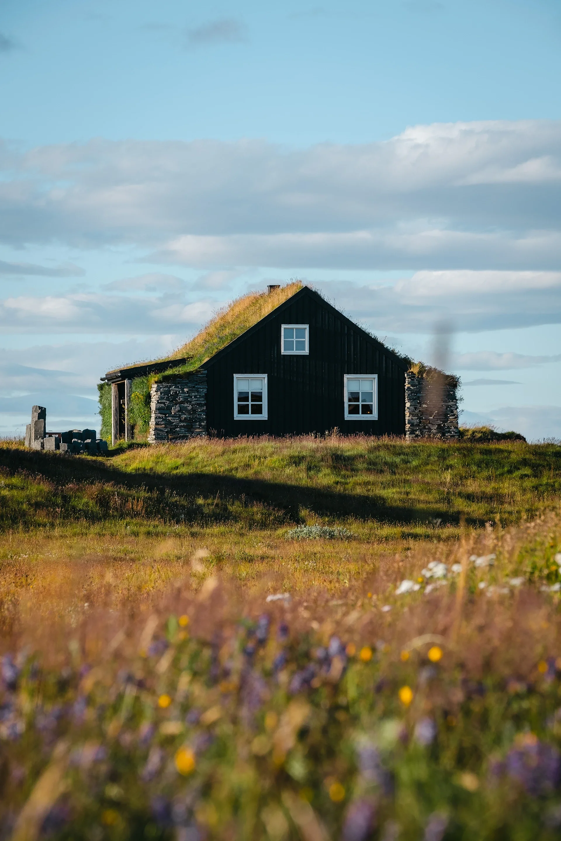 A black house with a grassy roof on a grassy field under a blue sky with clouds.