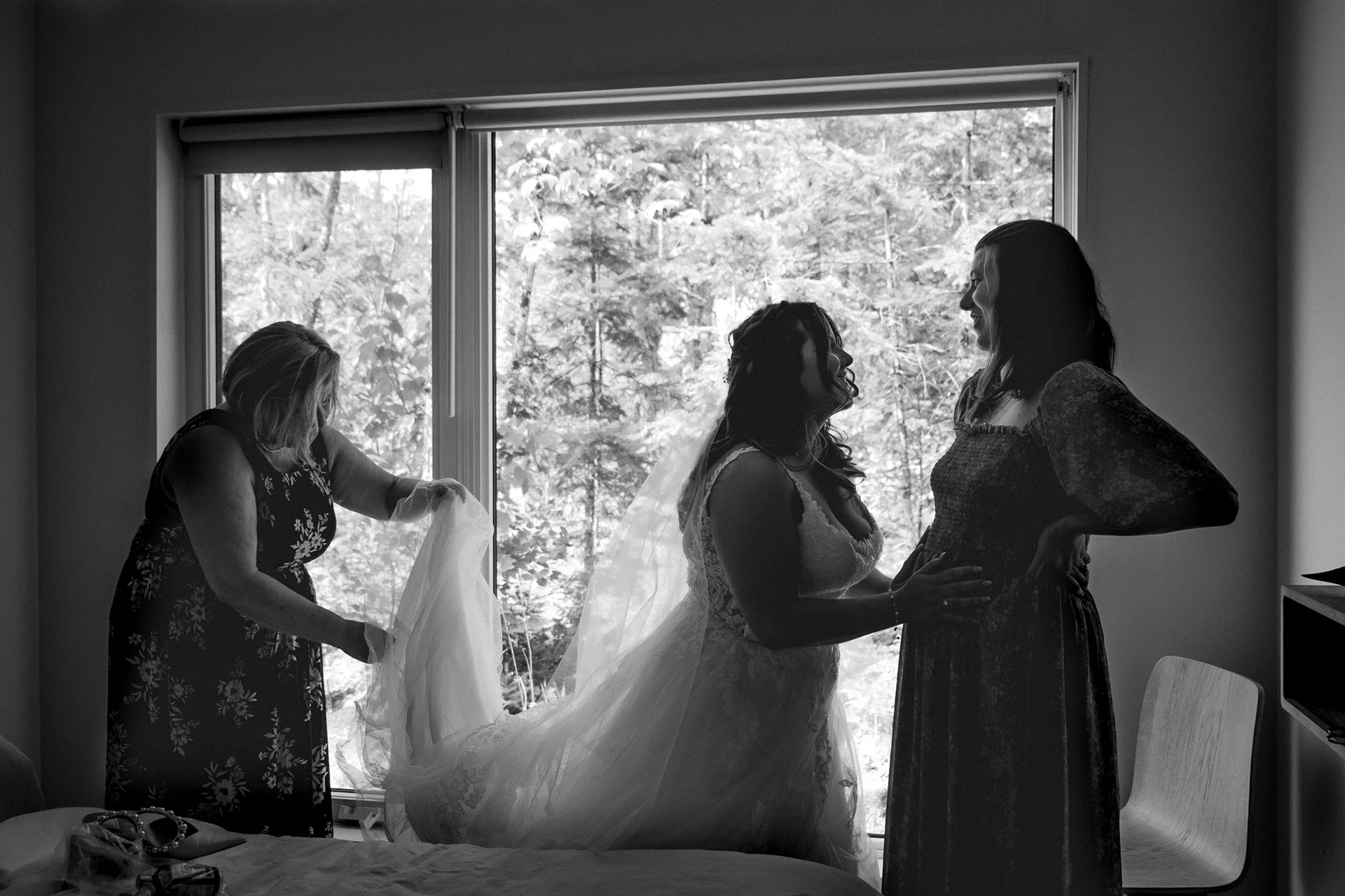 Three women preparing for a wedding, one in a wedding dress with veil, two others helping her, all silhouetted against a window with trees outside.