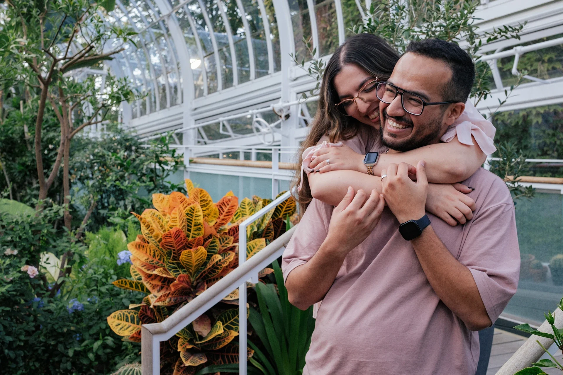 A couple sharing an embrace in a greenhouse surrounded by plants, with the woman hugging the man from behind and both smiling.