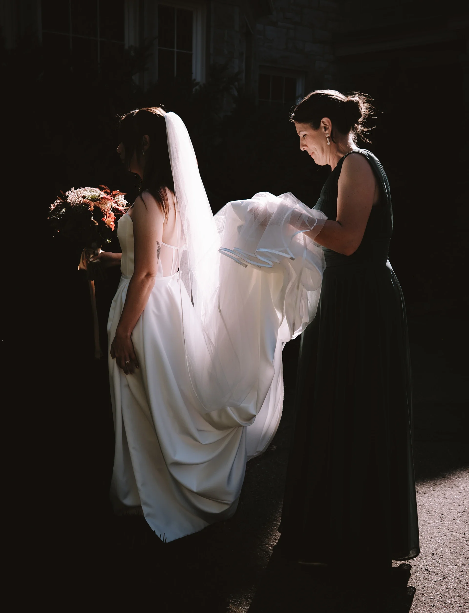 A bride in a wedding dress holding a bouquet and a woman in a black dress adjusting her dress outside in the sunlight.