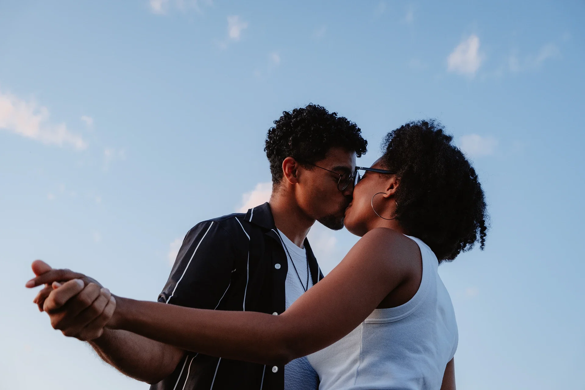 A couple kissing outdoors beneath a blue sky with scattered clouds.