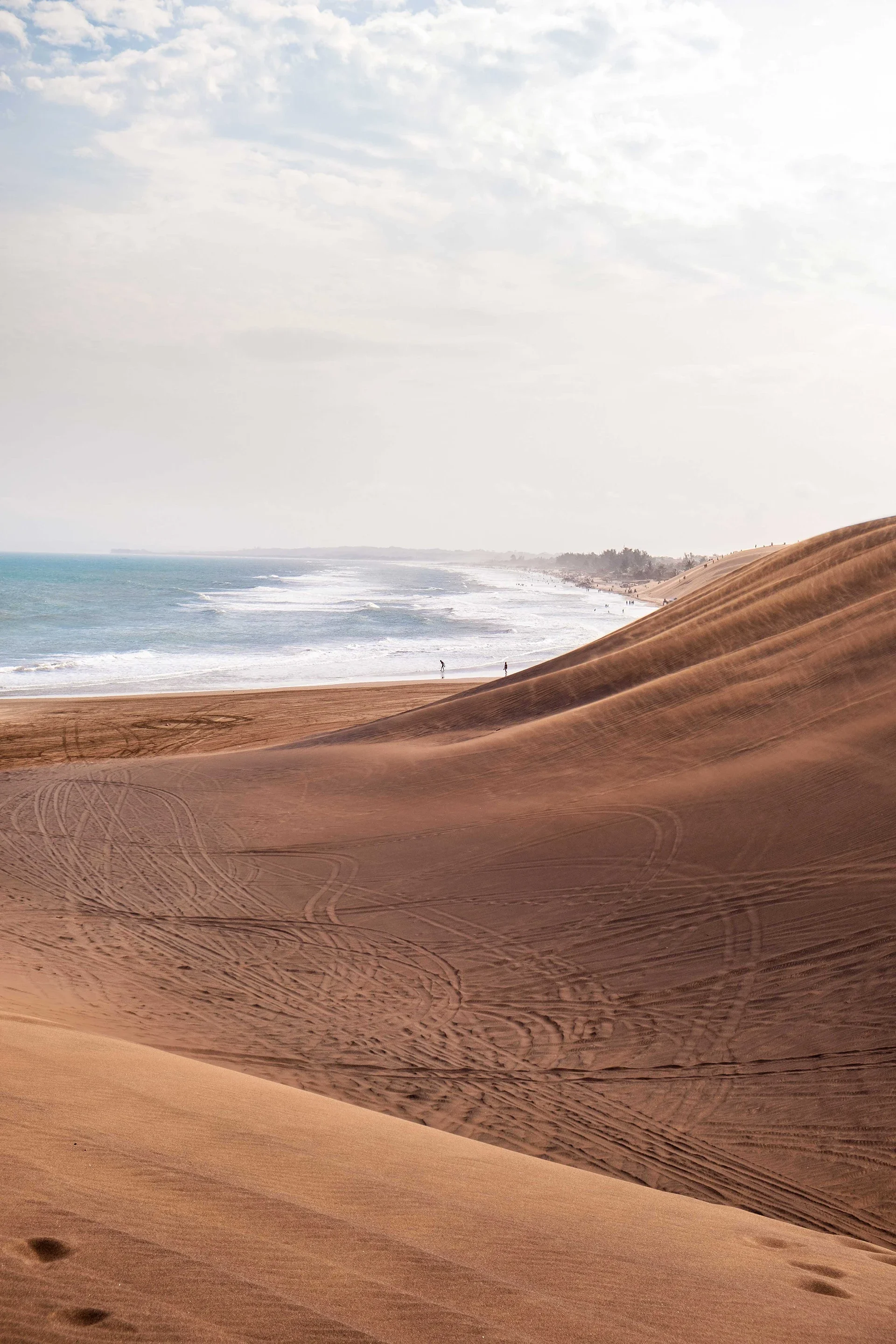 Sand dunes leading to a beach with ocean waves and cloudy sky in the background.