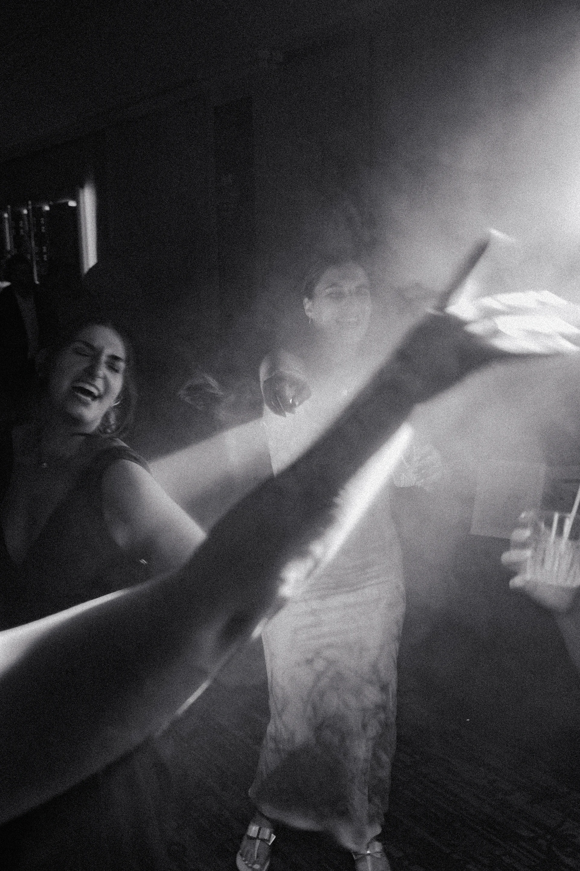 Black and white photo of three people at a party, smiling and laughing, with one person holding a drink. The scene has a hazy, celebratory atmosphere.