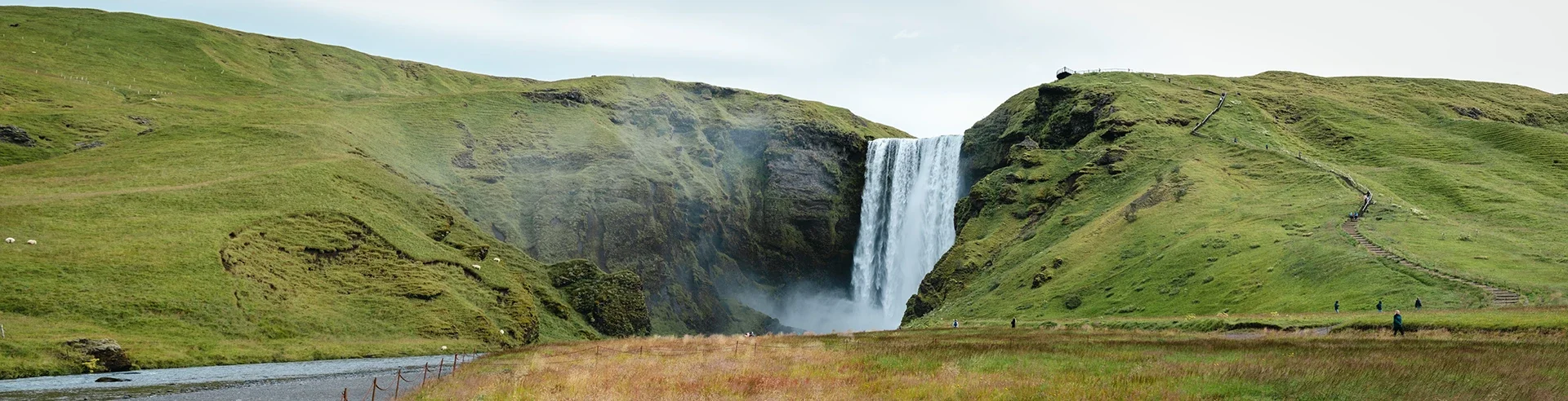 A scenic view of a waterfall cascading down a green hillside with a small river in the foreground and hiking stairs on the right side.