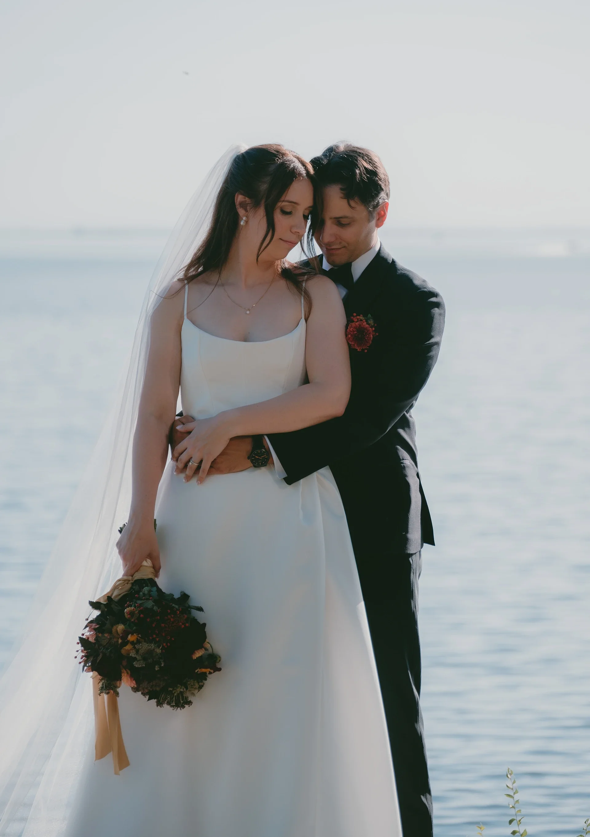 A bride and groom embrace on a beach, with the bride holding a bouquet of flowers and both looking tenderly at each other.
