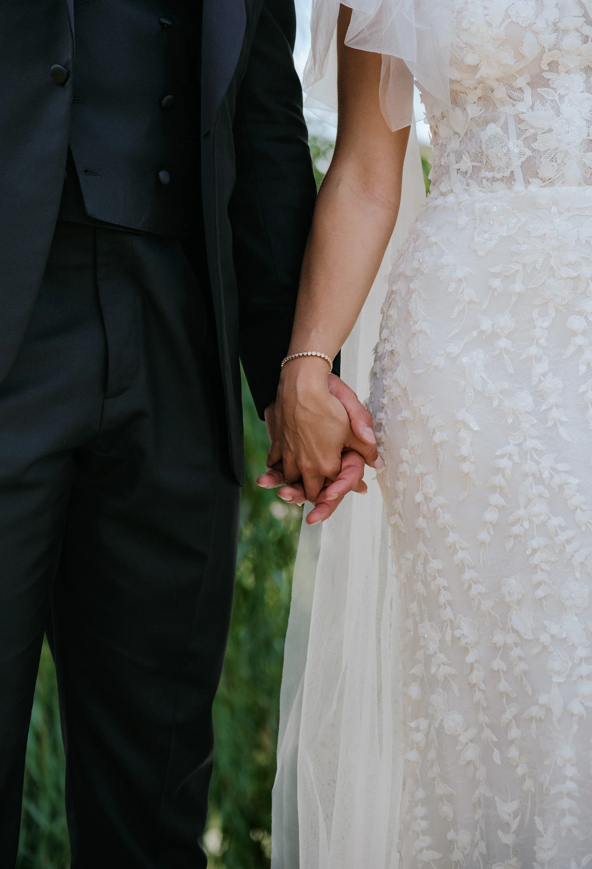 Close-up of a bride and groom holding hands during a wedding ceremony, with the bride wearing a lace wedding dress and the groom in a black suit.