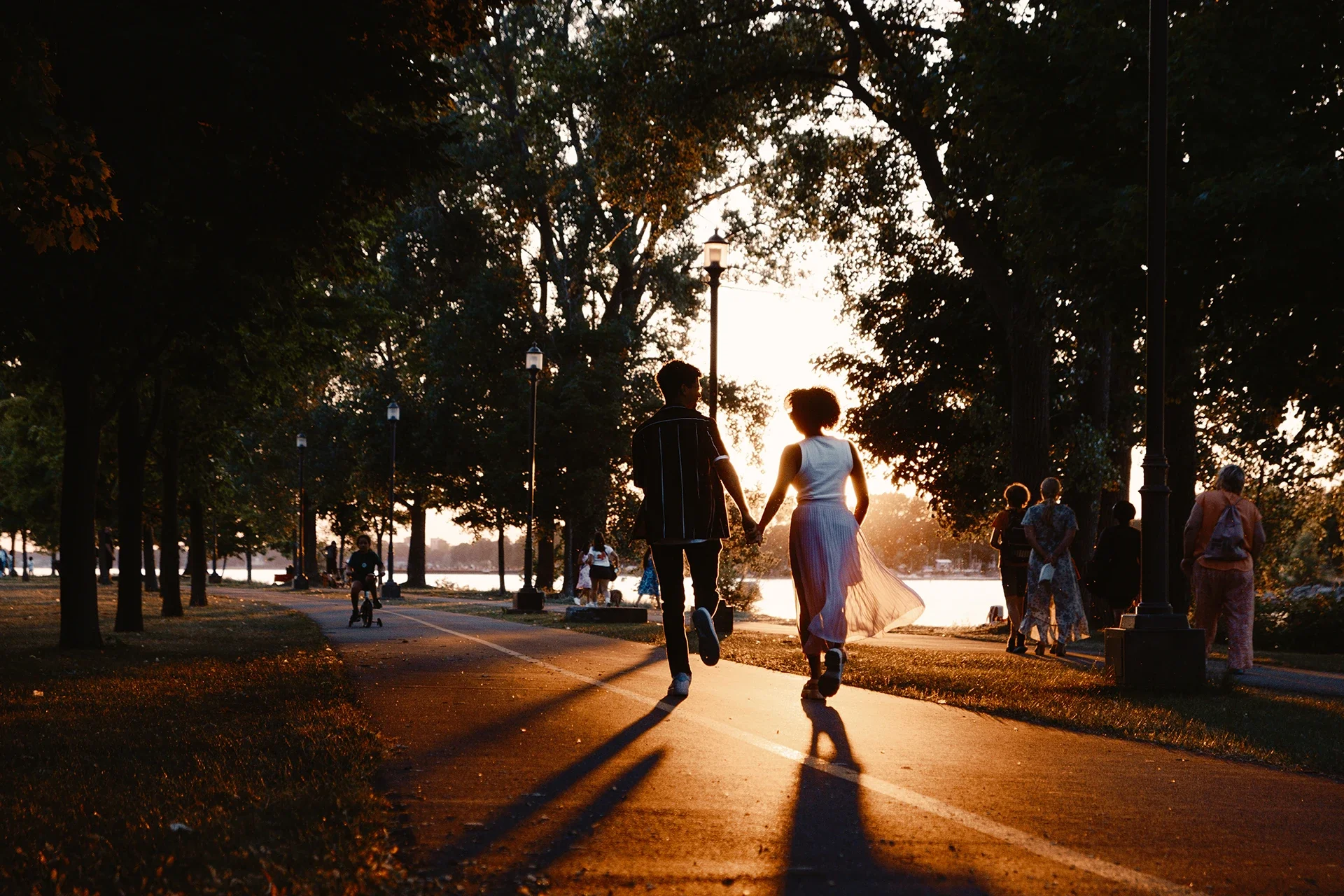 Couple holding hands and walking on a park pathway at sunset, with trees and other people in the background.
