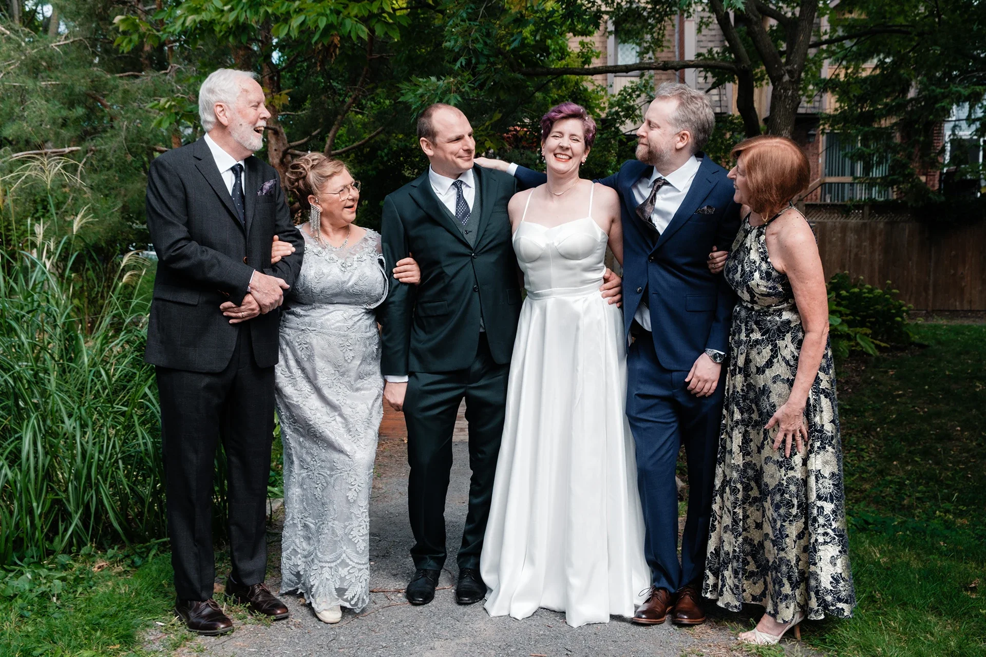 Group of six people at a wedding, including the bride in a white gown and five others in formal attire, standing outdoors among trees and greenery, smiling and interacting.