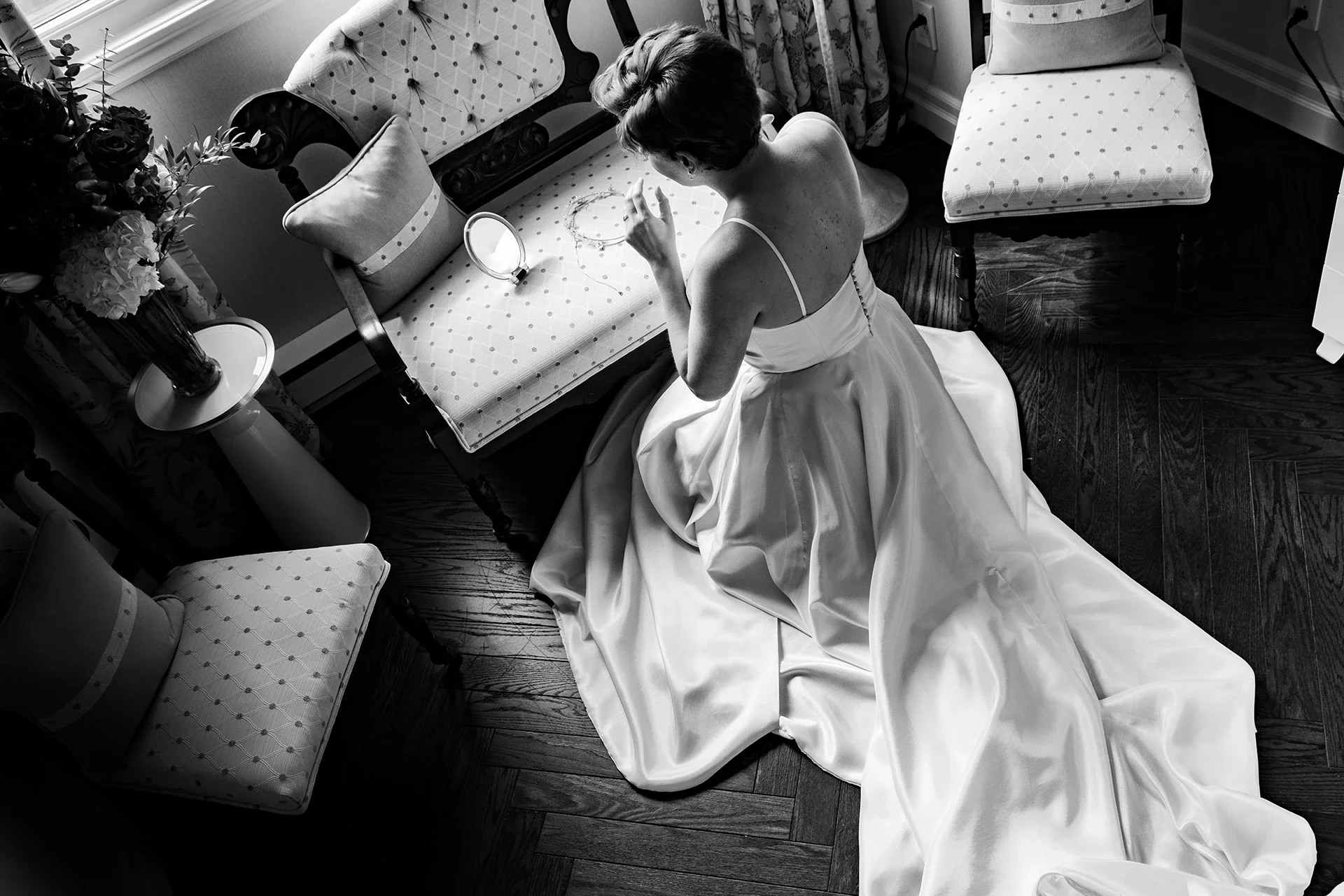 A woman in a white satin gown kneels on the floor, looking at herself in a mirror, in a room with hardwood floors, chairs, and a bouquet of flowers.