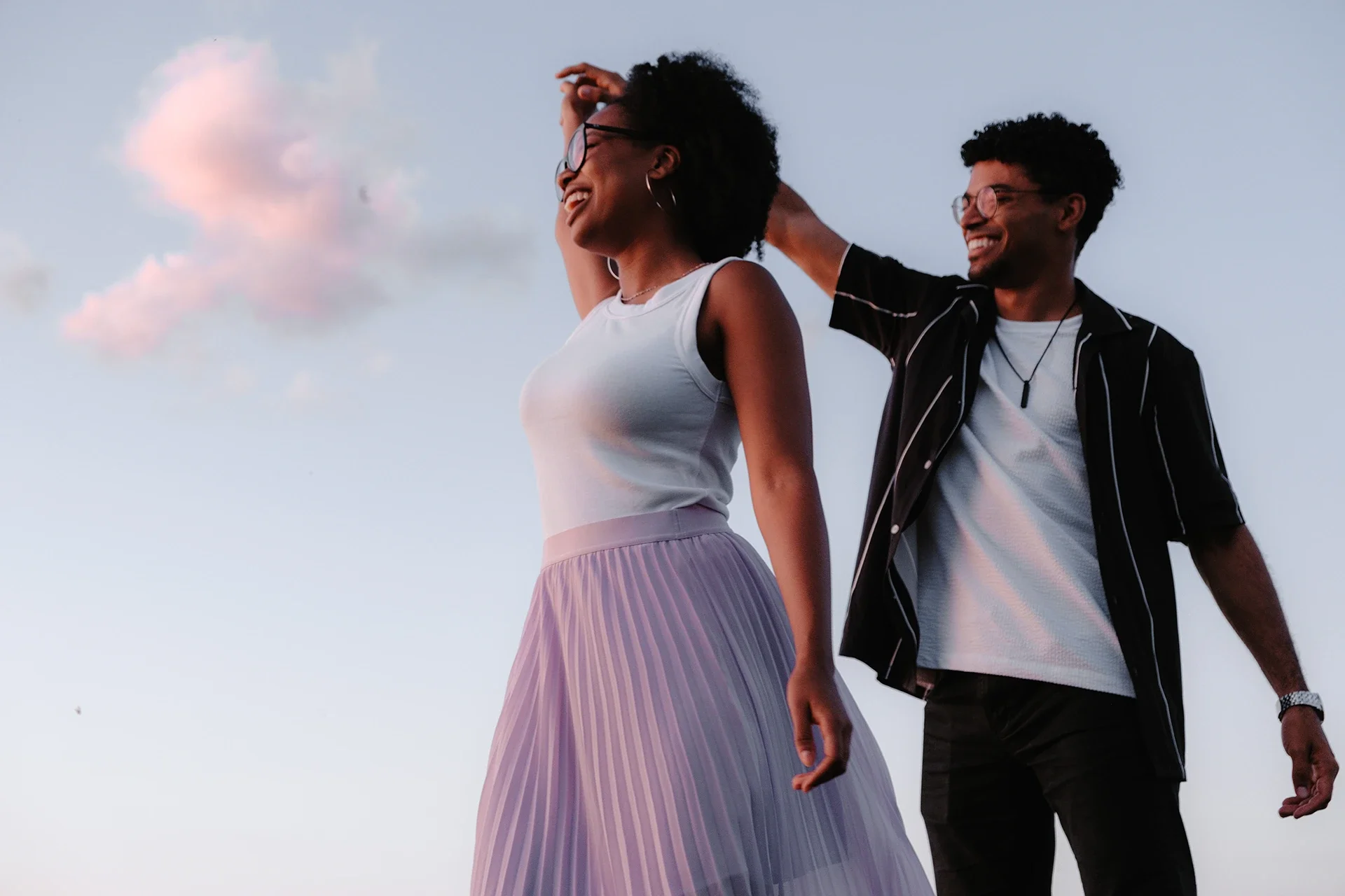 A smiling woman and man outside during sunset, with the woman wearing a white sleeveless top and a light purple pleated skirt, and the man wearing a black short-sleeve shirt with white stripes and glasses, appearing to enjoy each other's company.