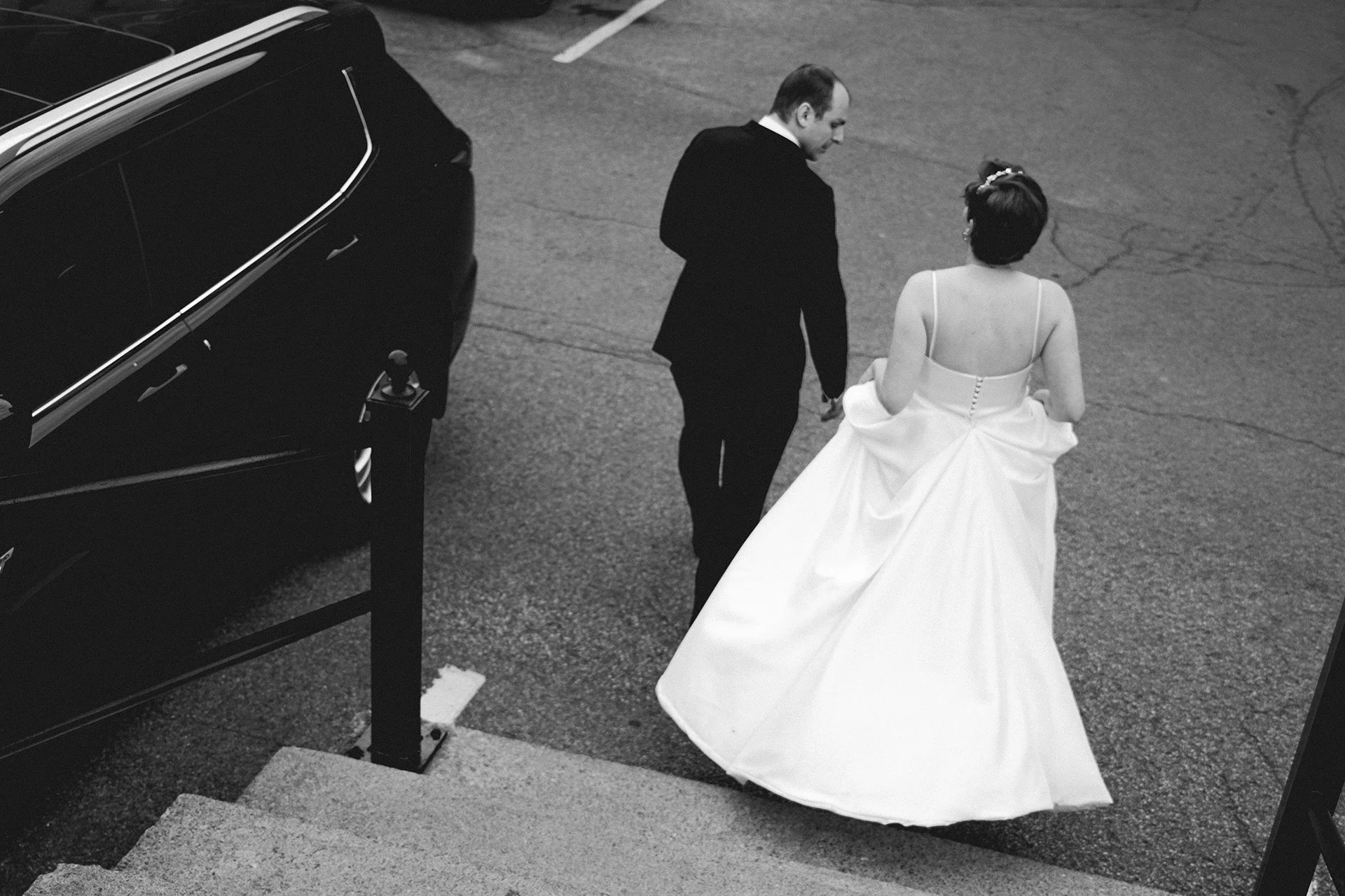 A black and white photo of a bride and groom walking together outside near stairs and a parked car, dressed in wedding attire.