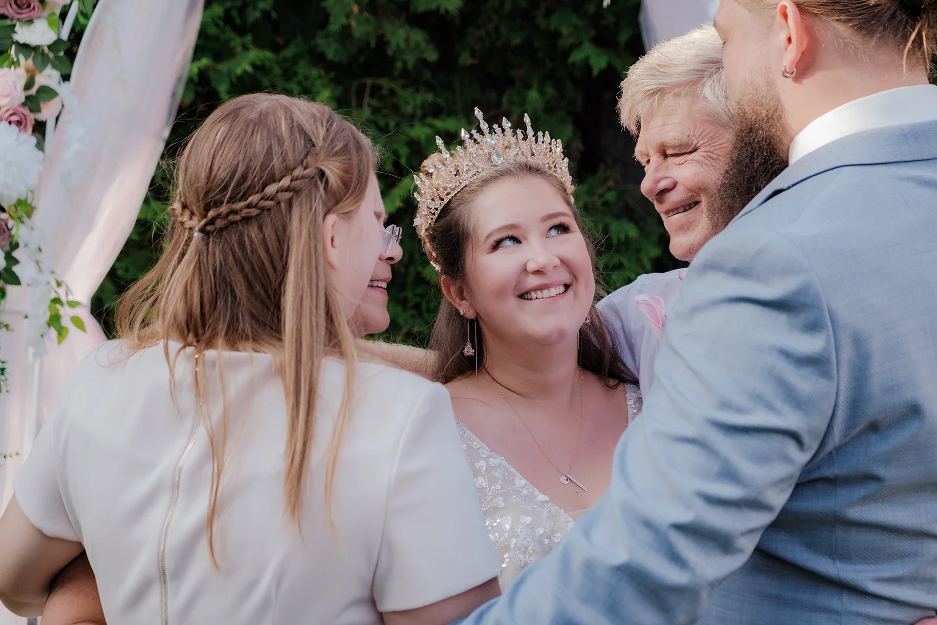 A wedding ceremony outdoors with a bride in a tiara and necklace smiling with three other people around her, including a man in a gray suit and a woman with glasses and a braid.