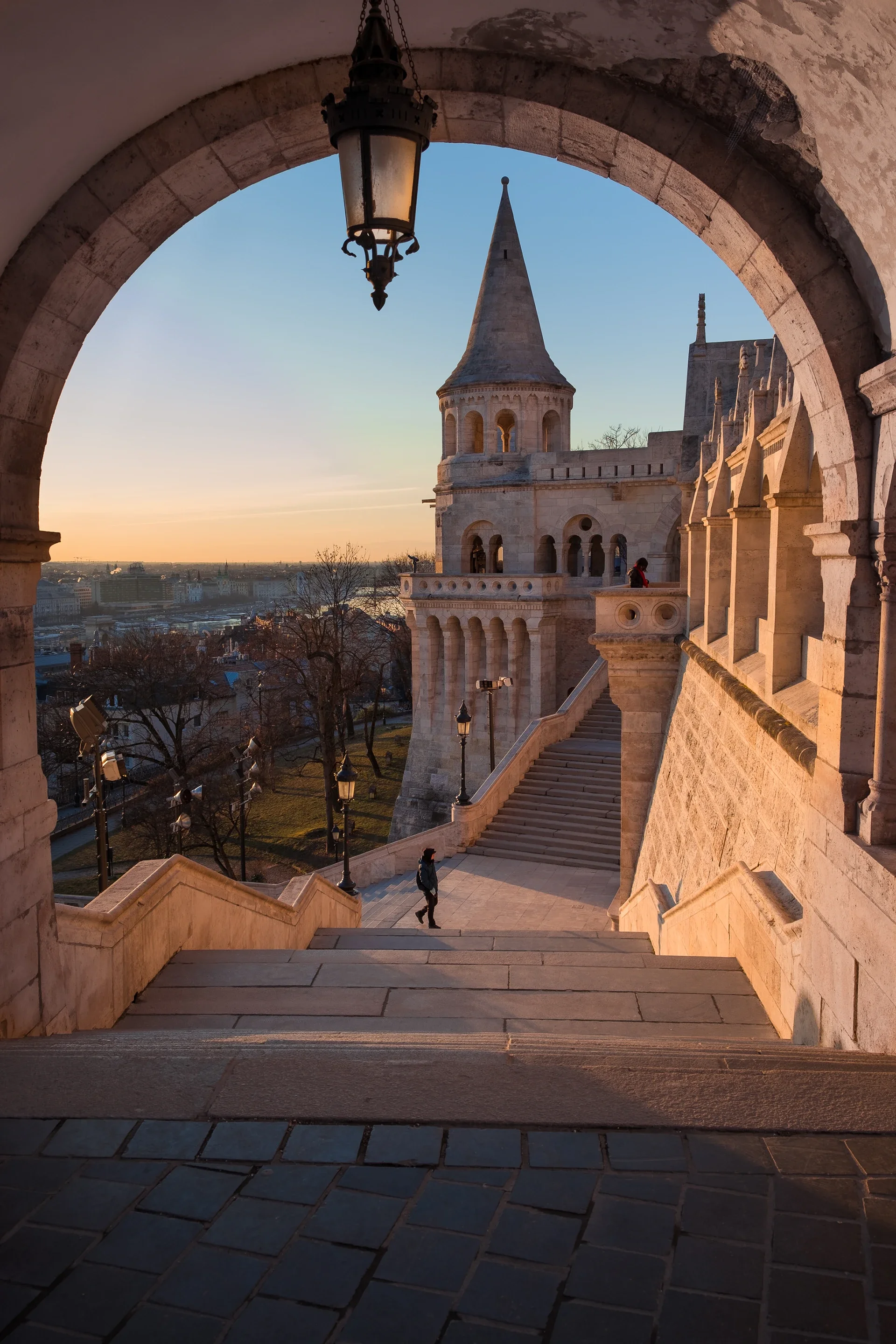 View of Fisherman's Bastion in Budapest at sunset, taken through an archway with a hanging lantern, featuring stairs leading down and a castle turret in the background.