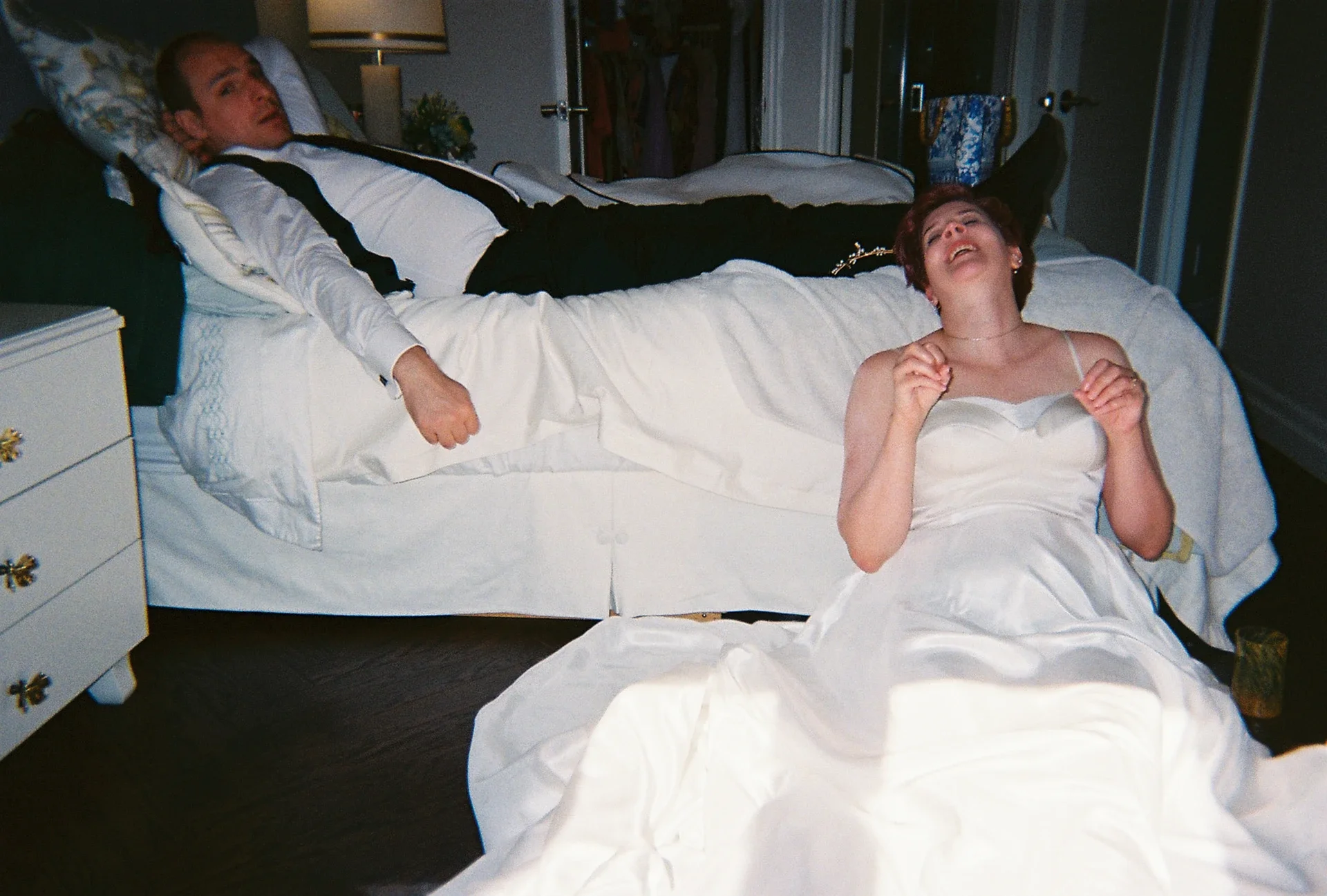 A man in a tuxedo lying on a bed and a woman in a wedding dress kneeling on the floor, both with distressed expressions.