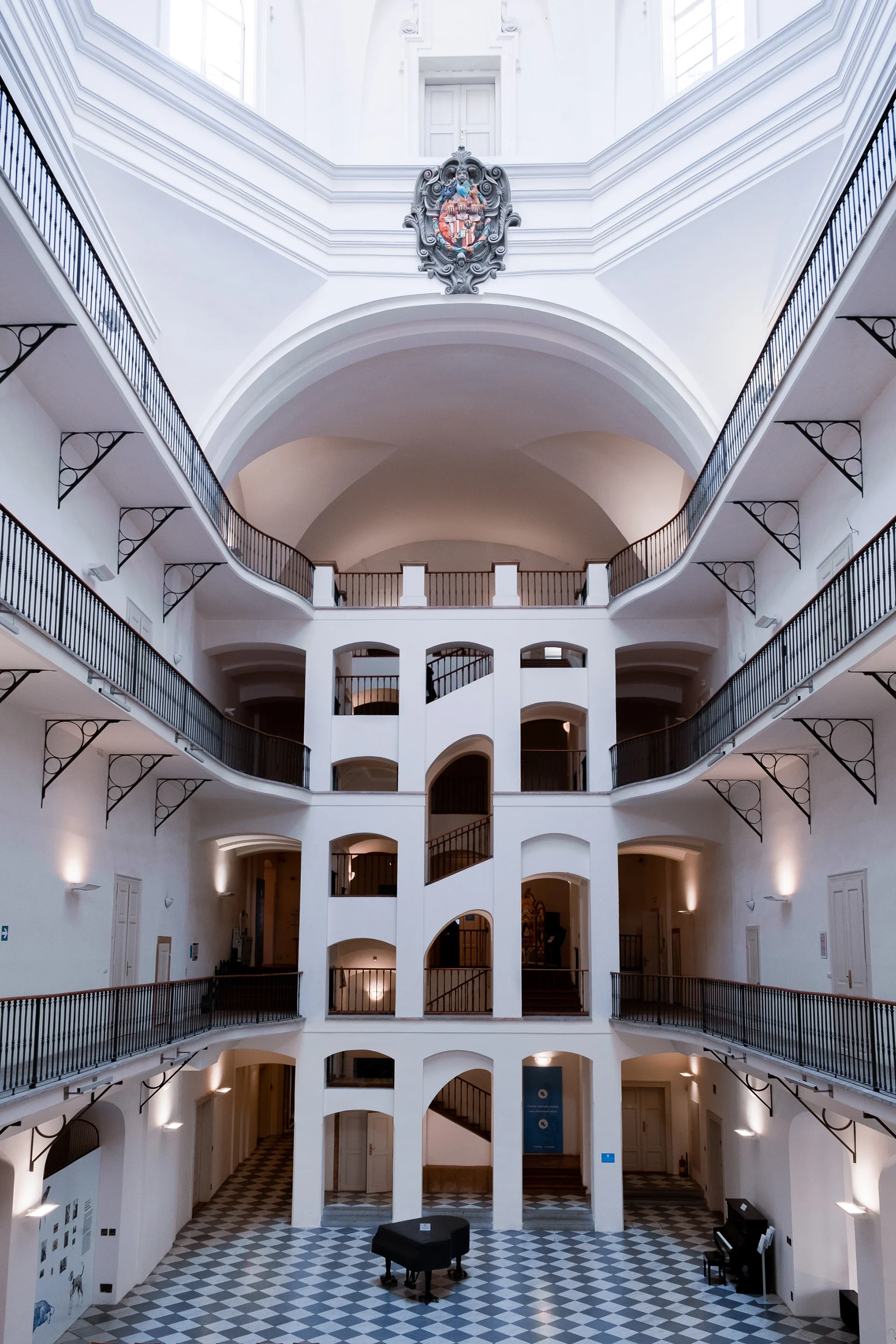 Interior of a multi-story building with white walls, arched openings, and a checkered black and white floor. There is a black grand piano on the ground level and a blue informational sign near the wall.