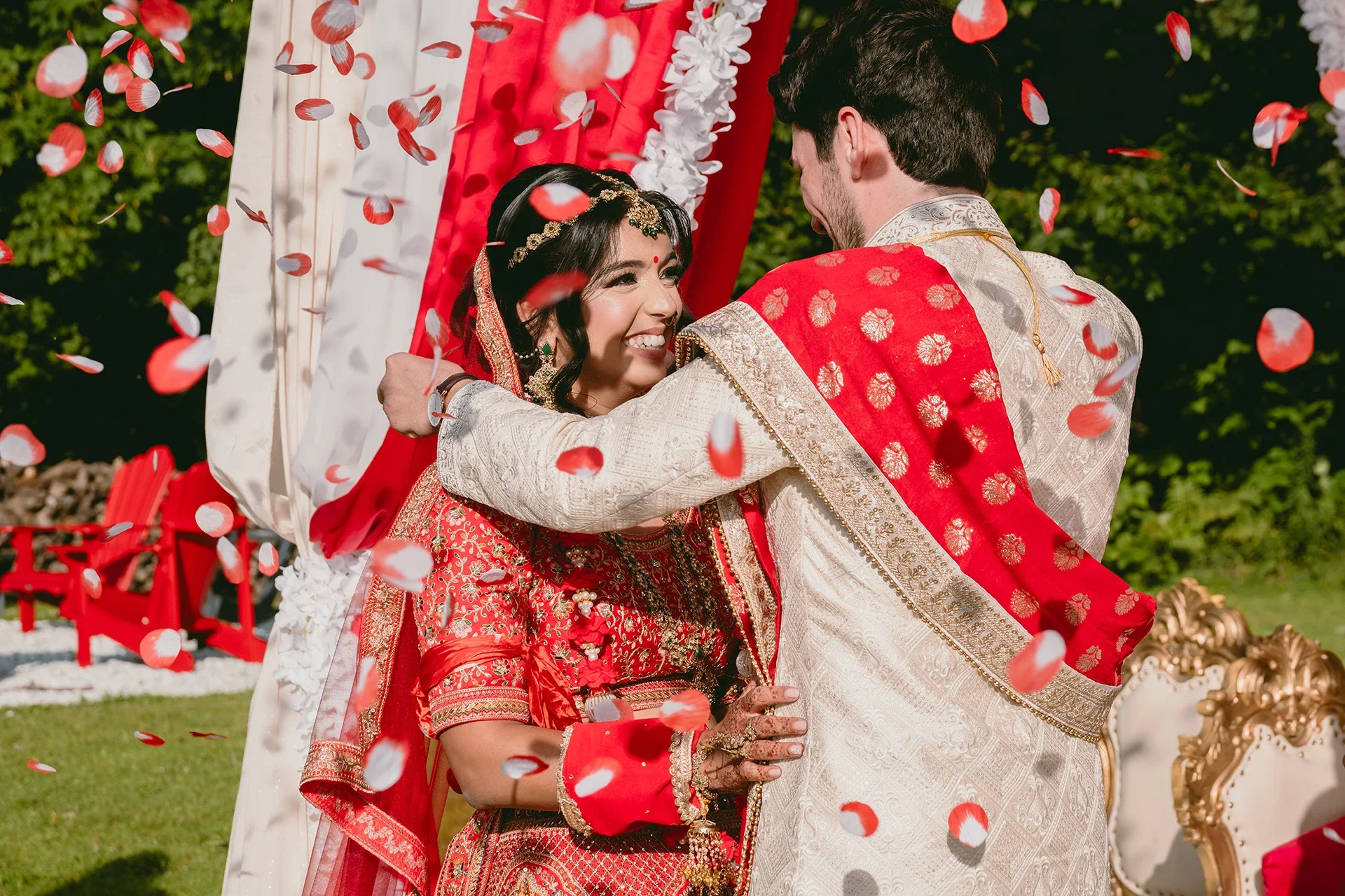 A bride and groom in traditional Indian wedding attire sharing a joyful moment while tossing flower petals outdoors.