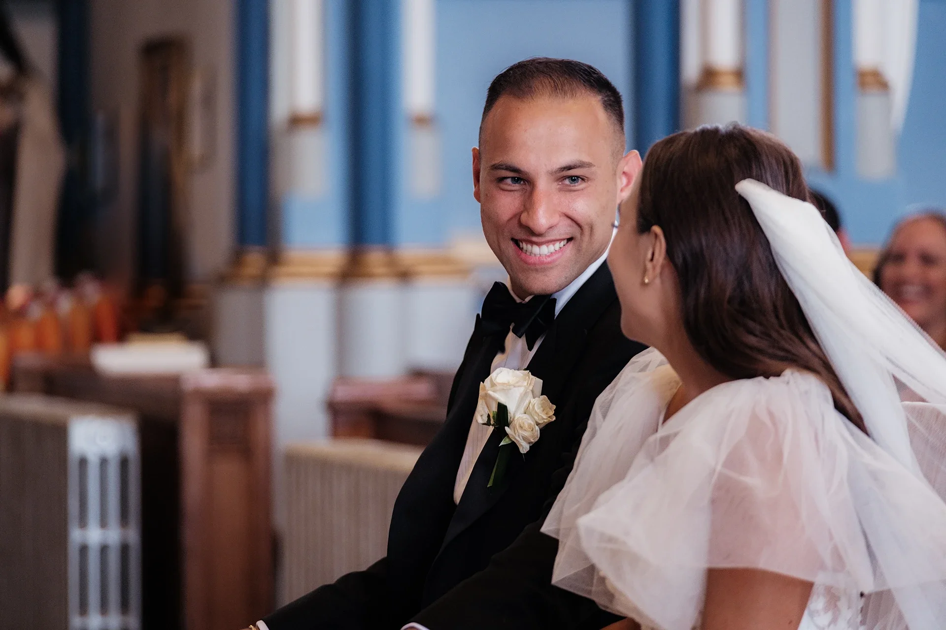 A bride and groom sitting together during a wedding ceremony inside a church, smiling at each other, with the groom wearing a tuxedo and the bride wearing a white dress and veil.