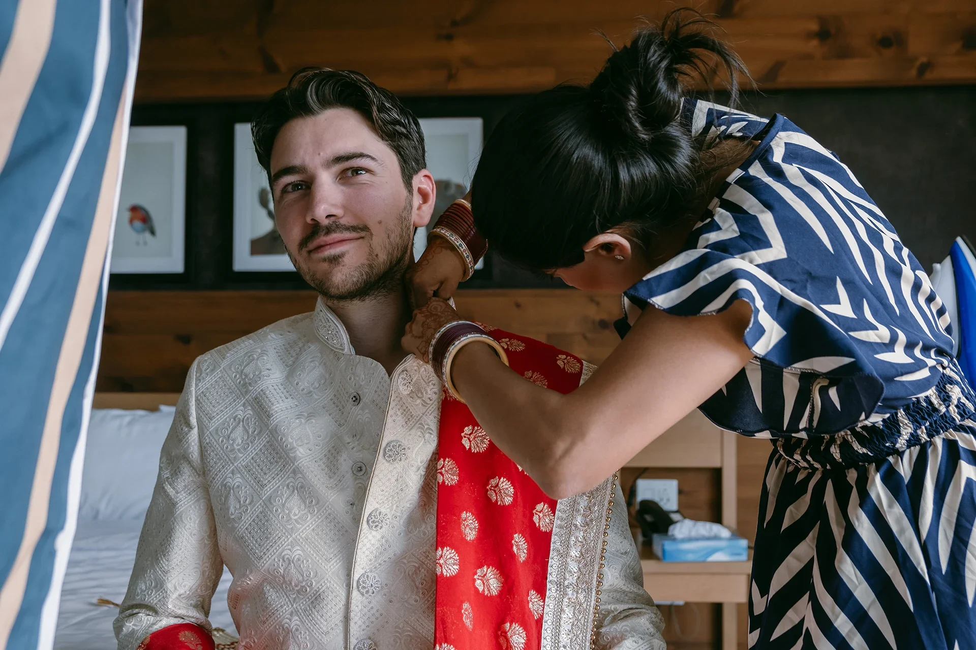 A man wearing traditional Indian wedding attire is sitting on a bed, while a woman in a striped dress is adjusting his red and gold embroidered shawl. The man has dark hair, a light beard, and is smiling gently. The woman has black hair tied back and