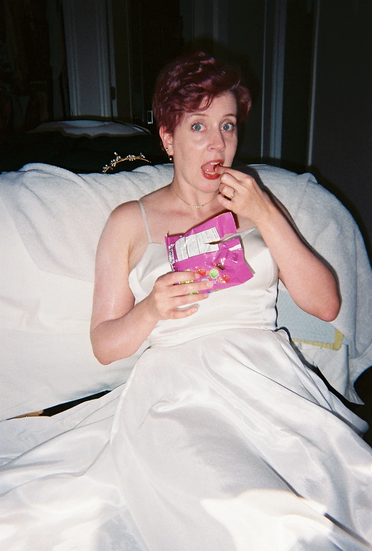 A woman dressed in a white satin gown sitting on a bed, eating candy from a pink snack bag, with a surprised expression.