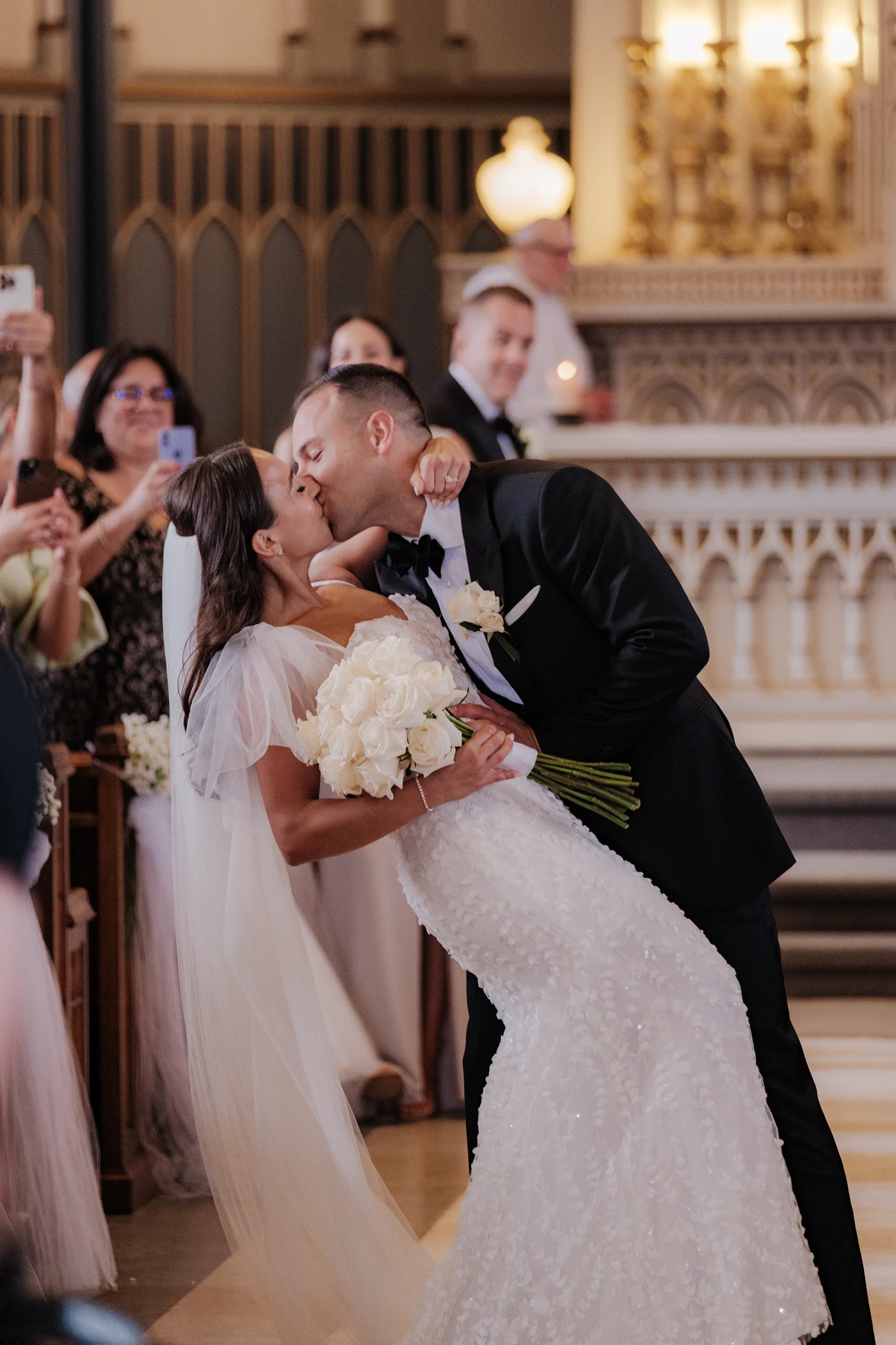 A newlywed couple shares a kiss during their wedding ceremony inside a church, surrounded by friends and family.