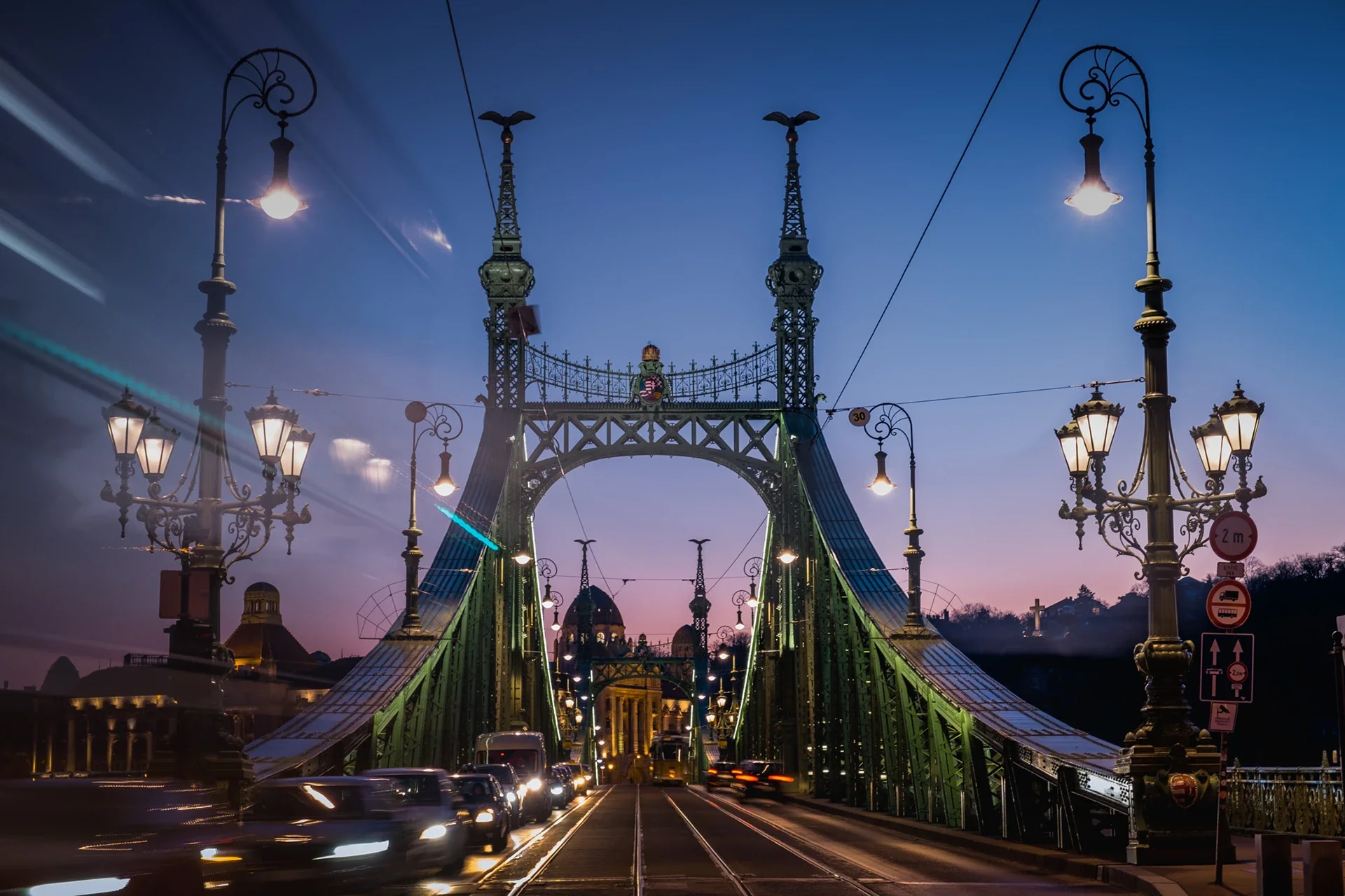 The Liberty Bridge in Budapest at dusk, illuminated with street lamps and traffic, with a clear sky in the background.