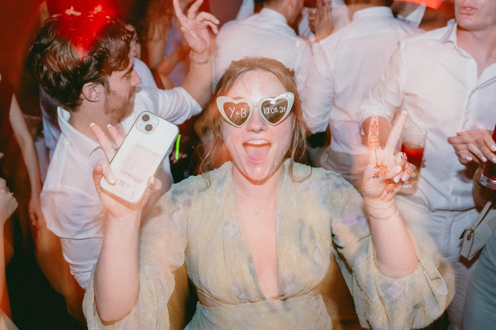A woman at a party wearing heart-shaped sunglasses with 'Y+B' and a date written on them, holding a smartphone, smiling, and making a peace sign with her hands.