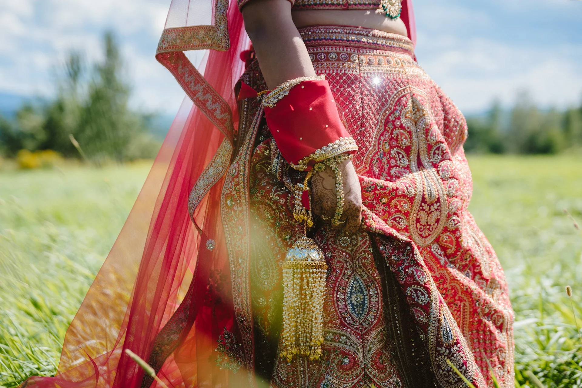 A woman dressed in traditional Indian wedding attire, holding a decorative umbrella, standing in a green field with a clear blue sky.