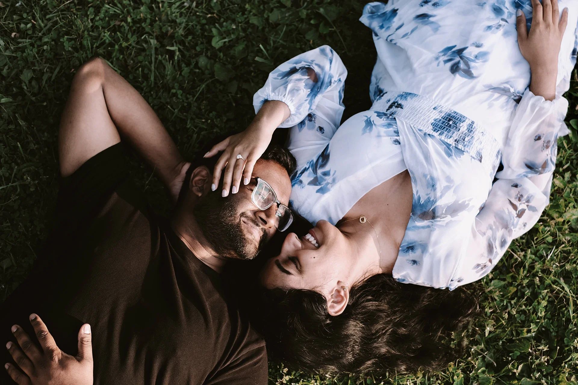 A man and woman lie on grass, facing each other, smiling and touching foreheads. The man wears a brown shirt and glasses, the woman wears a white and blue dress and gold jewelry.