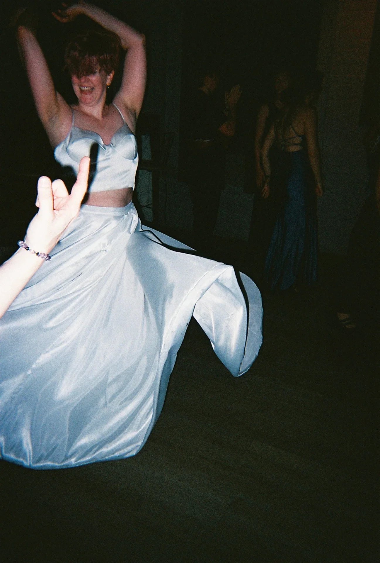 A woman in a shiny white satin dress is dancing with her arms raised and a big smile. She appears to be enjoying herself at a party or celebration, with other people in the background.