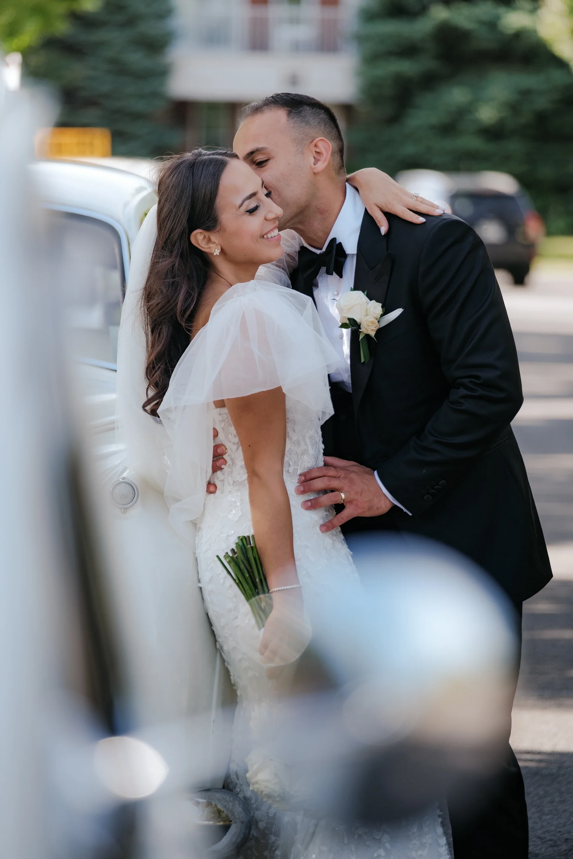 A newlywed couple sharing a kiss in front of a vintage car, with the groom in a tuxedo and the bride in a white wedding dress holding a bouquet.