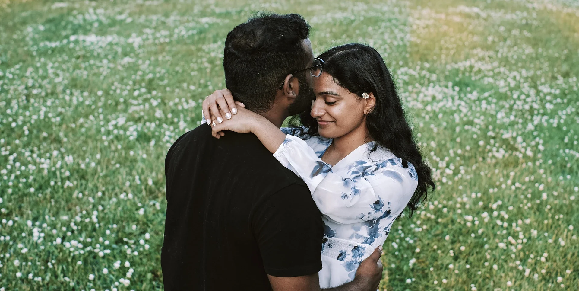 A couple embracing in a grassy field filled with small white flowers, with the woman smiling softly and the man wearing glasses.