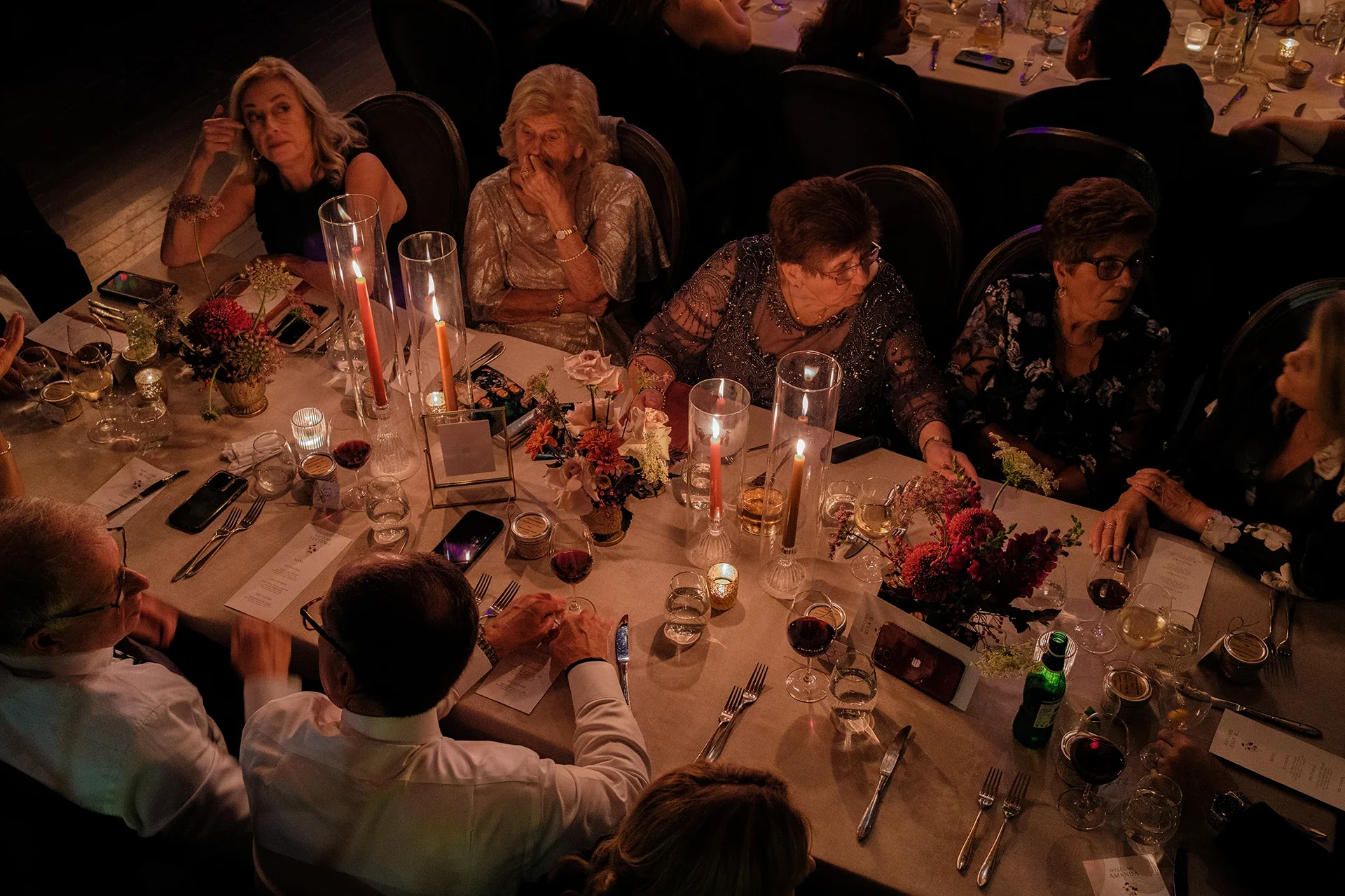 People sitting at a decorated dinner table with flowers, candles, and drinks in a dimly lit setting, possibly at a formal event.