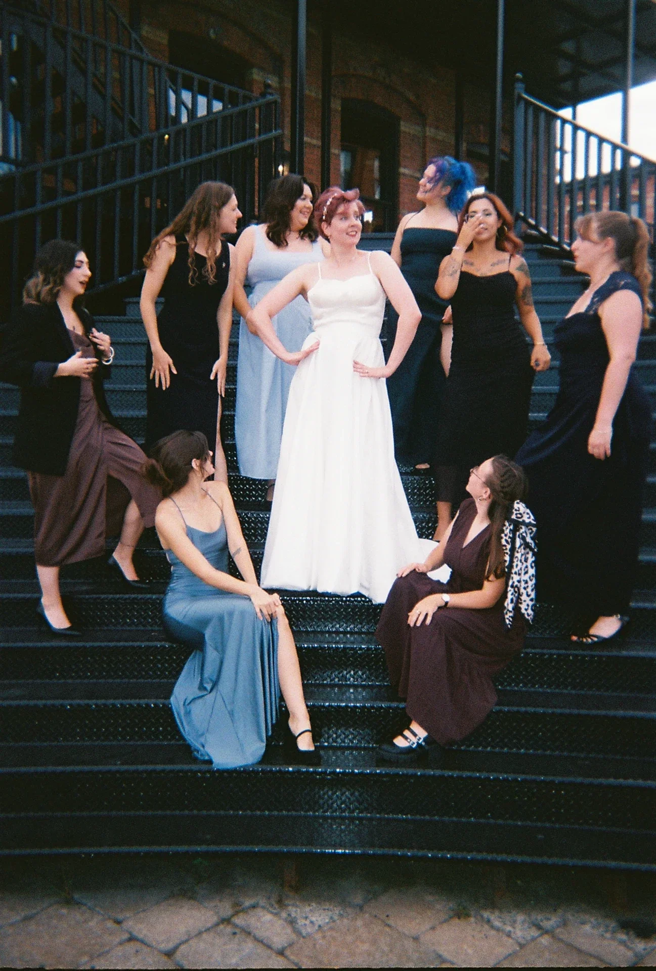A group of women dressed in formal dresses gathering on a staircase, with the woman in a white dress at the center, surrounded by women with varied hairstyles and colors, some sitting and some standing, engaging in conversation.