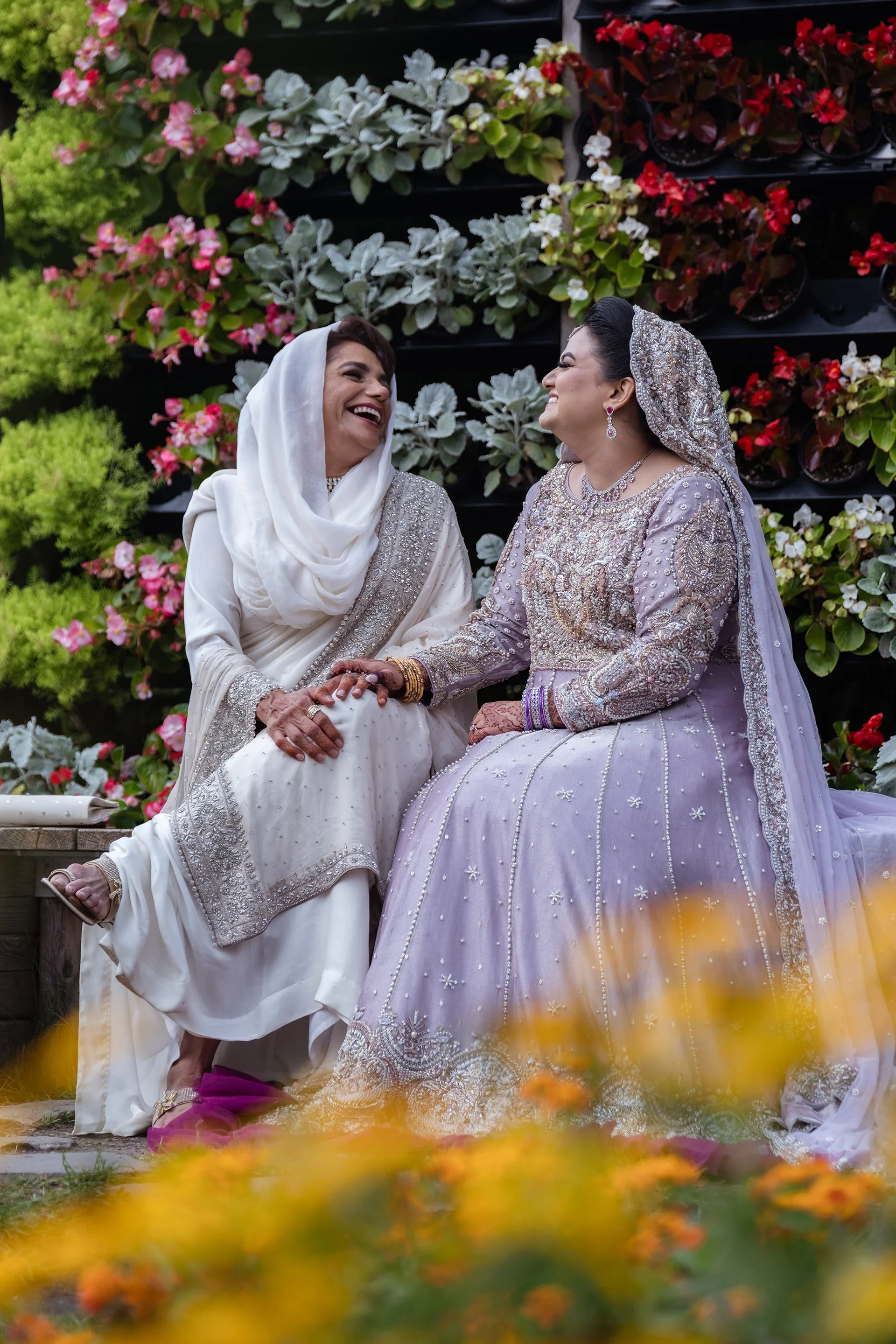 Two women dressed in traditional South Asian bridal attire sitting and smiling at each other in front of a floral backdrop.