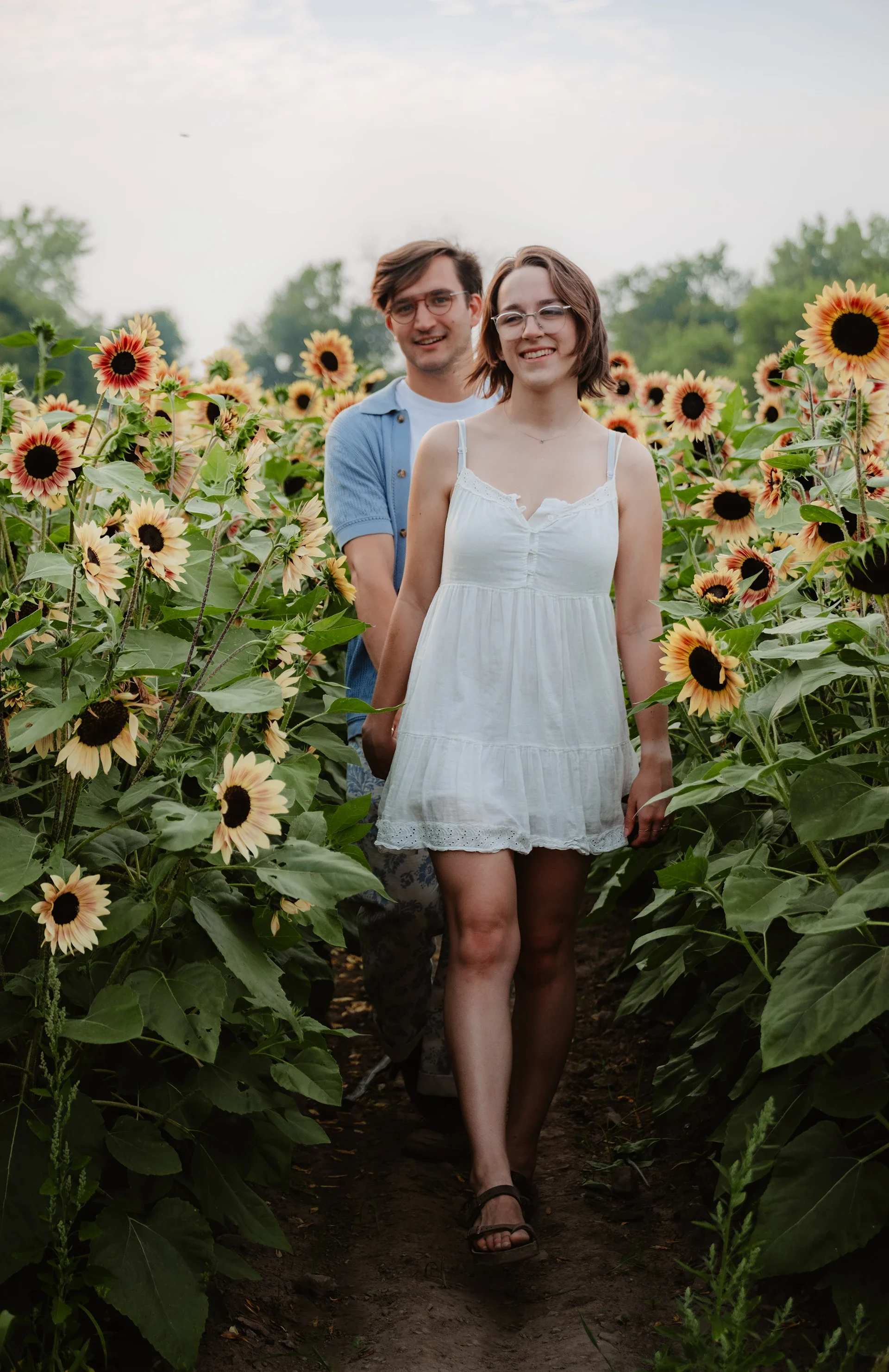 A young couple walking through a sunflower field on a cloudy day, smiling and holding hands.