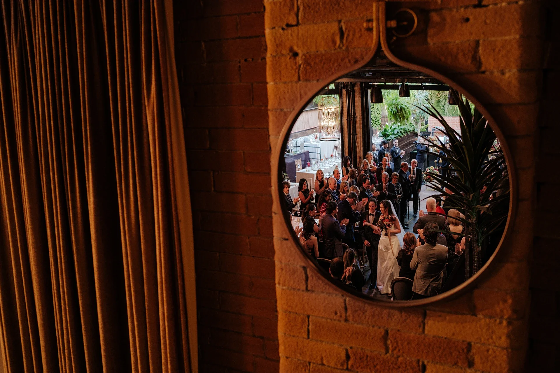 A wedding celebration viewed through a circular mirror on a brick wall. The bride and groom are surrounded by guests inside a decorated venue with lush greenery and elegant lighting.