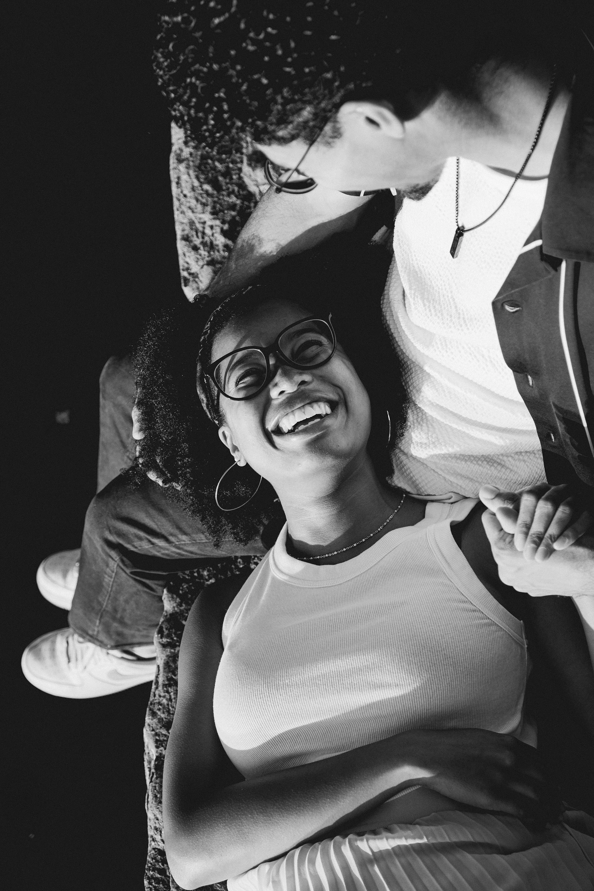 A black-and-white photo of two young people lying on the ground outdoors, smiling and looking at each other. One is a woman with glasses and hoop earrings, the other is a man with a beard wearing glasses and a necklace.