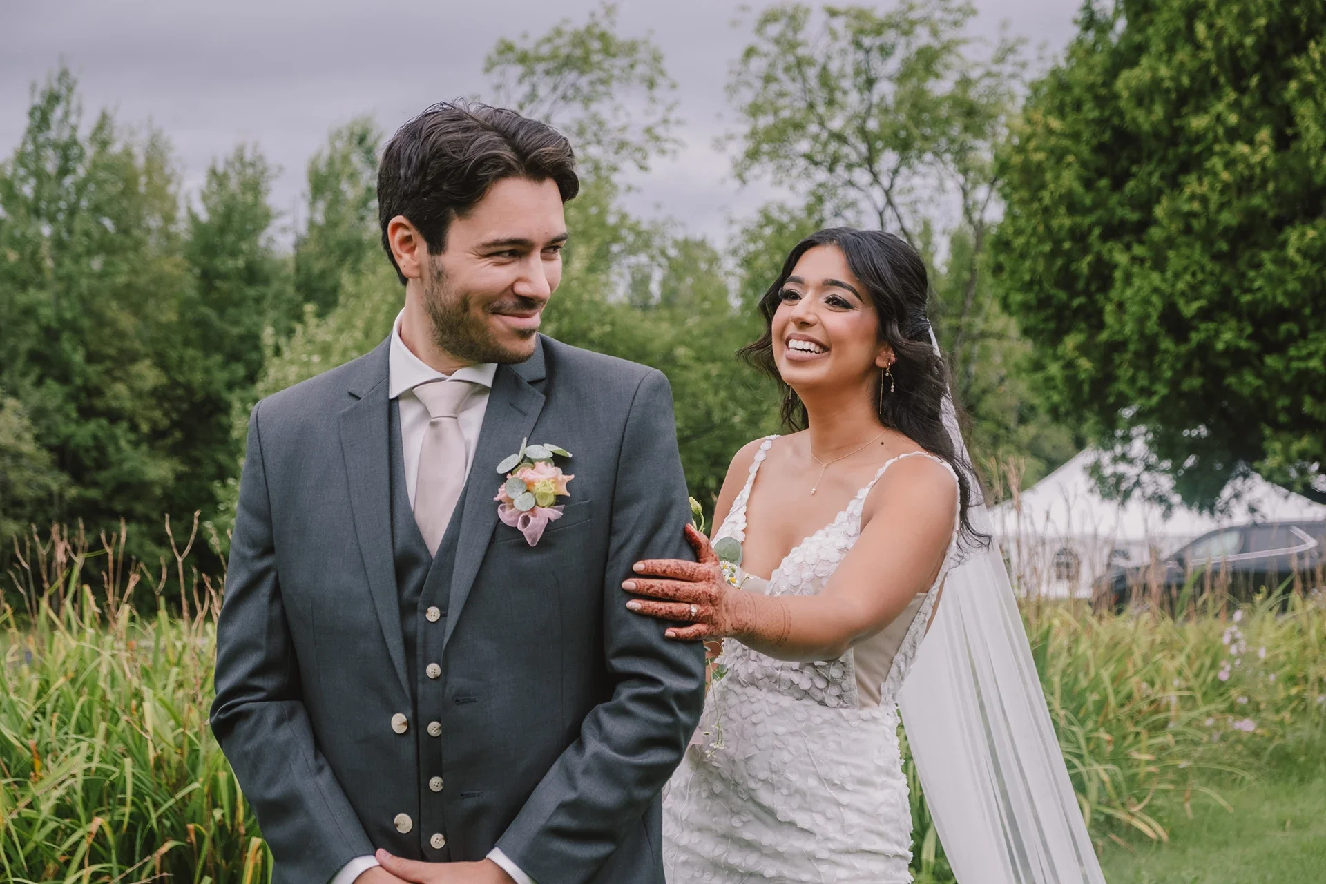 A bride and groom standing outdoors on their wedding day, smiling at each other. The bride has henna on her hands and is touching the groom's arm, wearing a white dress with a veil, while the groom is dressed in a gray suit with a boutonniere.