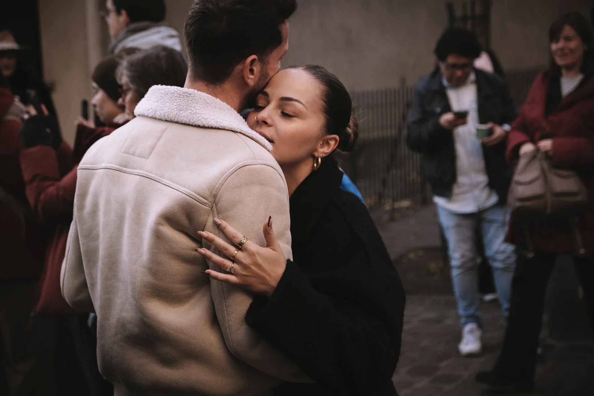 A couple embraces closely, with the woman resting her head on the man's shoulder. They are outdoors, and a group of people is visible in the background, some using their phones.
