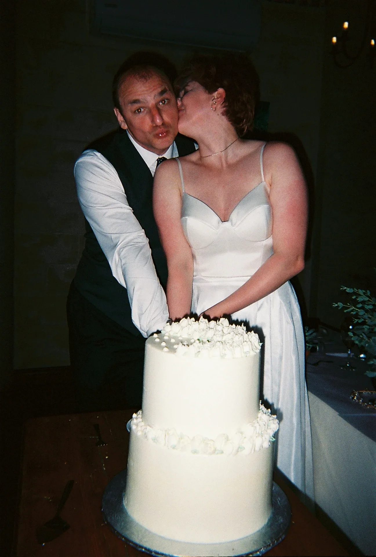 A couple, dressed in wedding attire, cutting a white wedding cake together, with a woman kissing the man on the cheek.