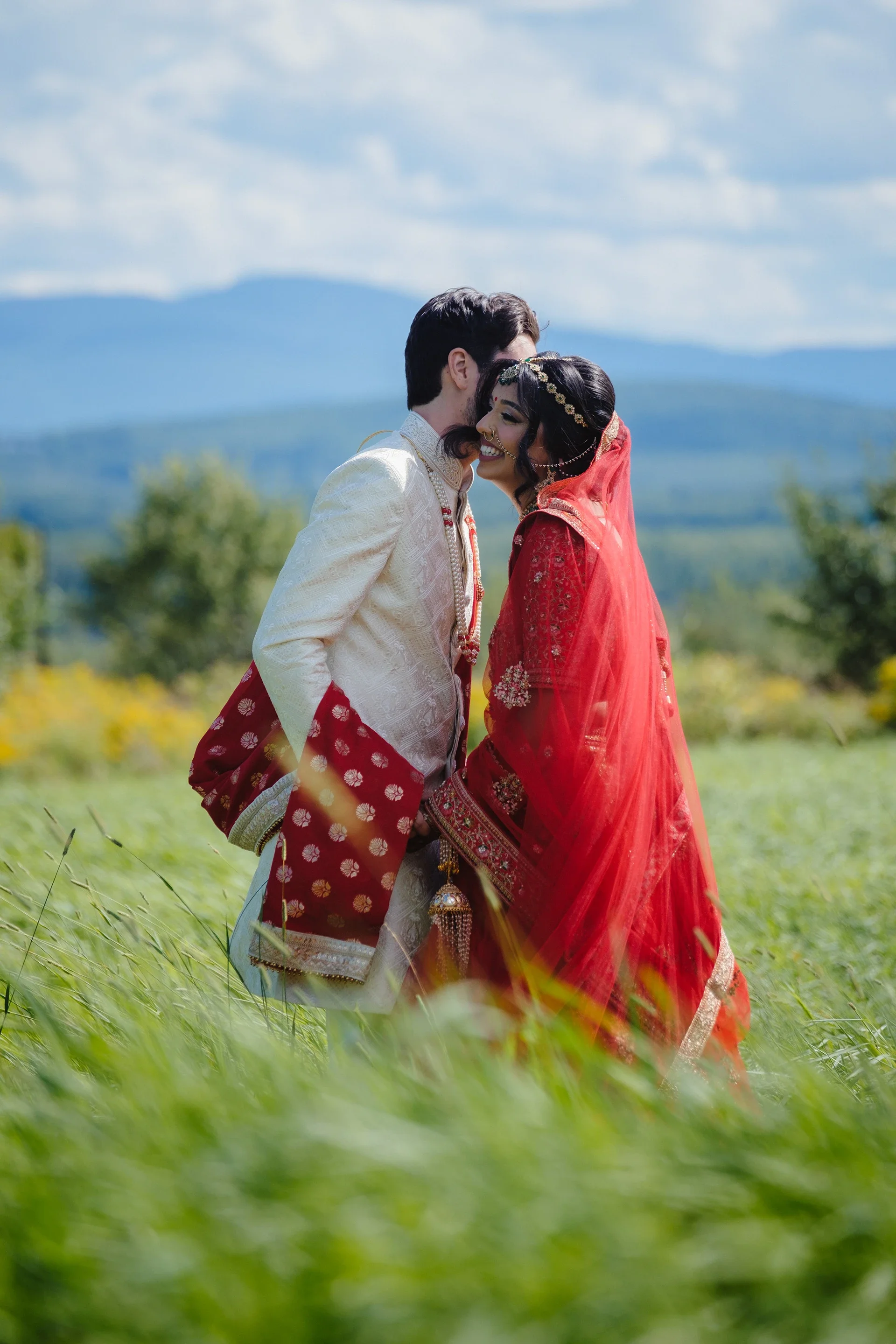 A happy Indian bride and groom in traditional attire standing outdoors in a green field with mountains in the background, sharing an affectionate moment.