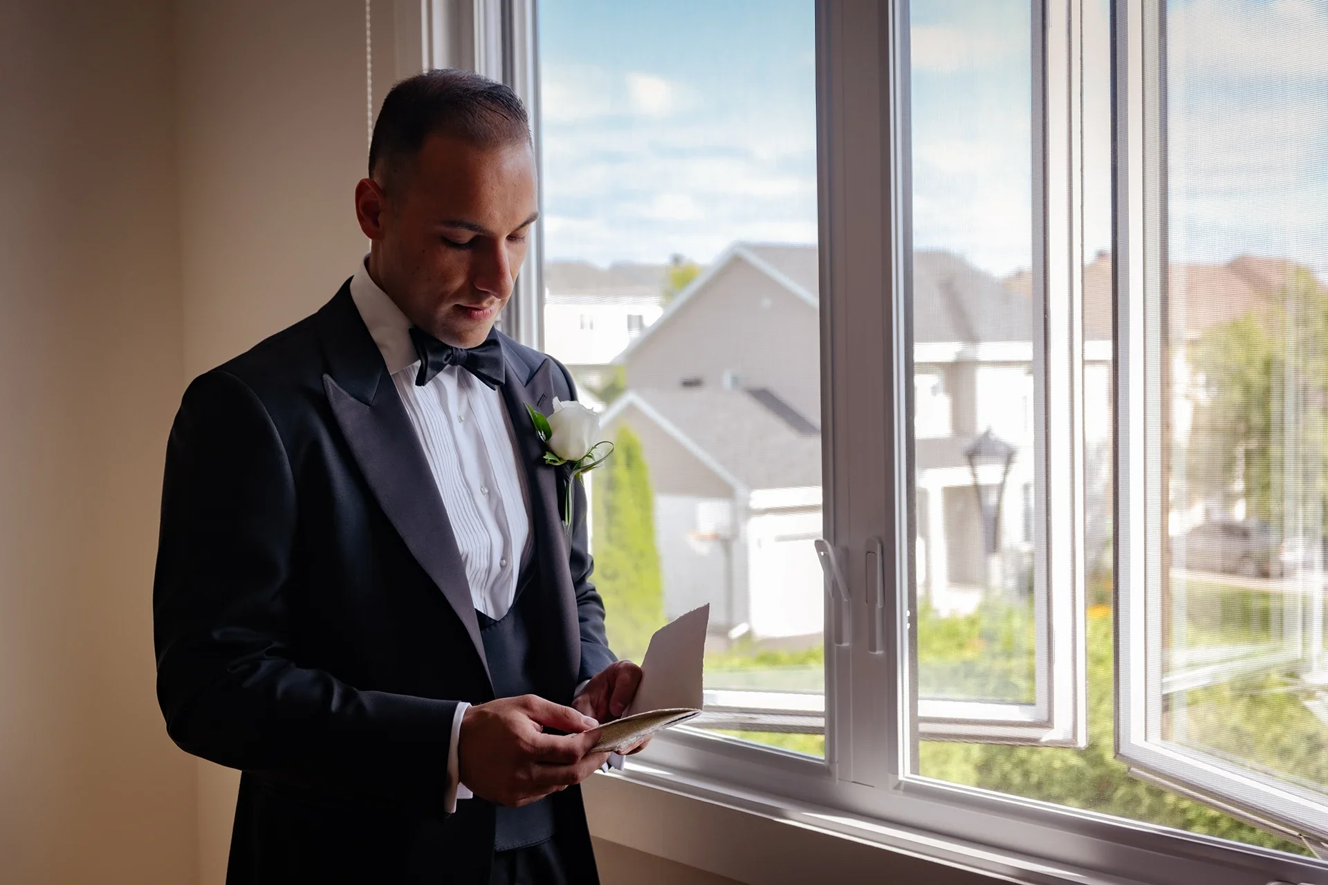 A man in a tuxedo reading a card by a window.