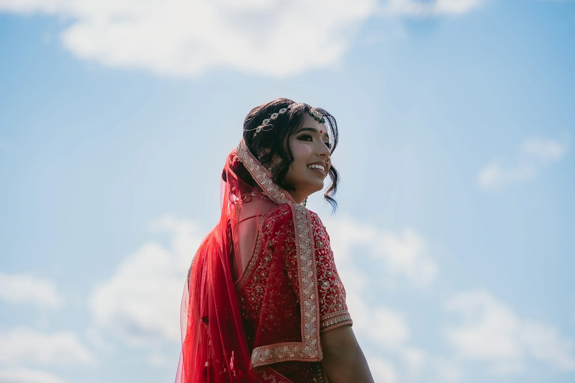 A smiling woman wearing a red traditional dress with intricate embroidery and a sheer red shawl, standing outdoors against a blue sky with scattered white clouds.