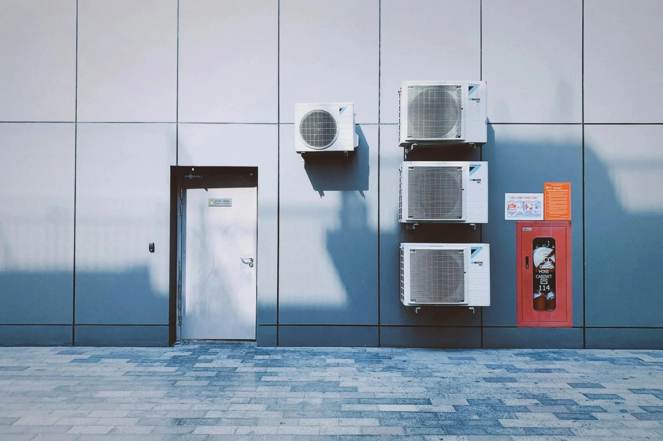 Exterior wall with four air conditioning units mounted vertically, a metallic door, and a red fire hose cabinet, with a paved ground.