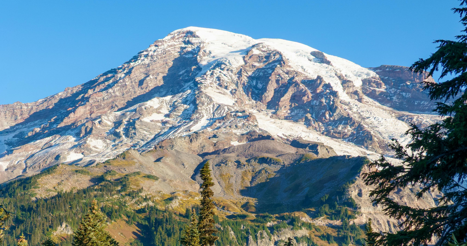Snow-covered mountain with rocky slopes and green forest in the foreground, clear blue sky