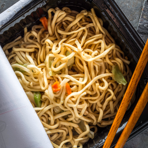 A black plastic takeout container filled with cooked ramen noodles, along with small pieces of green and orange vegetables, and a pair of yellow chopsticks resting on the side.